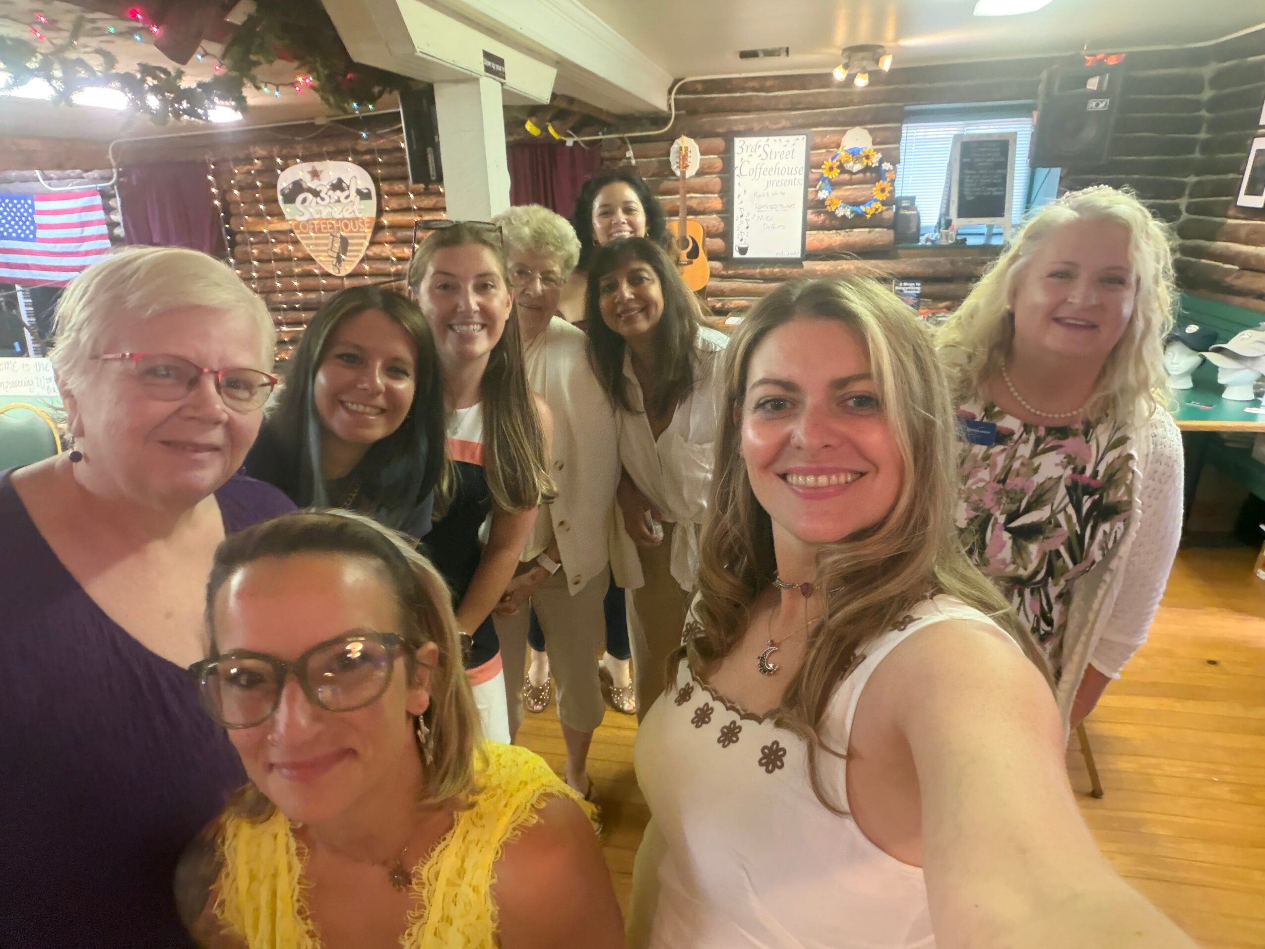 Group of eight women smiling in a room with brick walls and decorations, some wearing glasses, one taking a selfie.