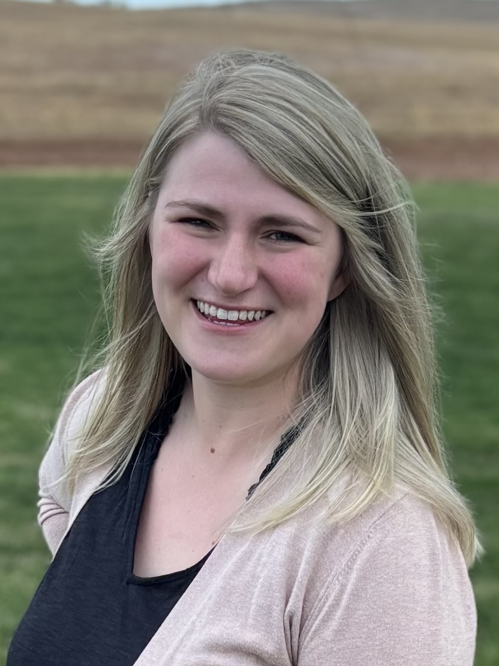 Smiling woman with blonde hair outdoors, green grass and field in background