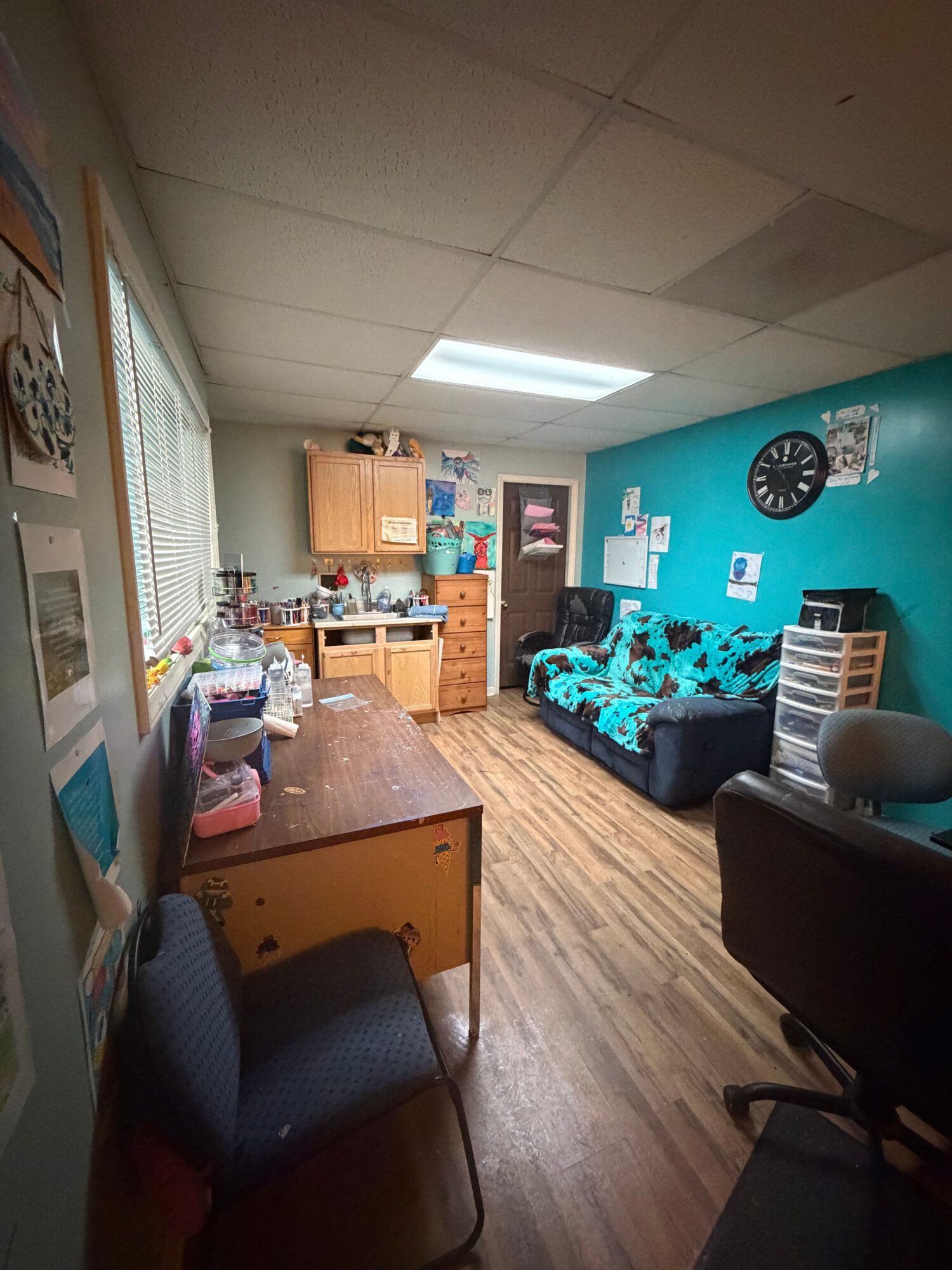 Office room with a dartboard, bookshelf, desk, chair, and a bed with blue bedding, viewed from the side.