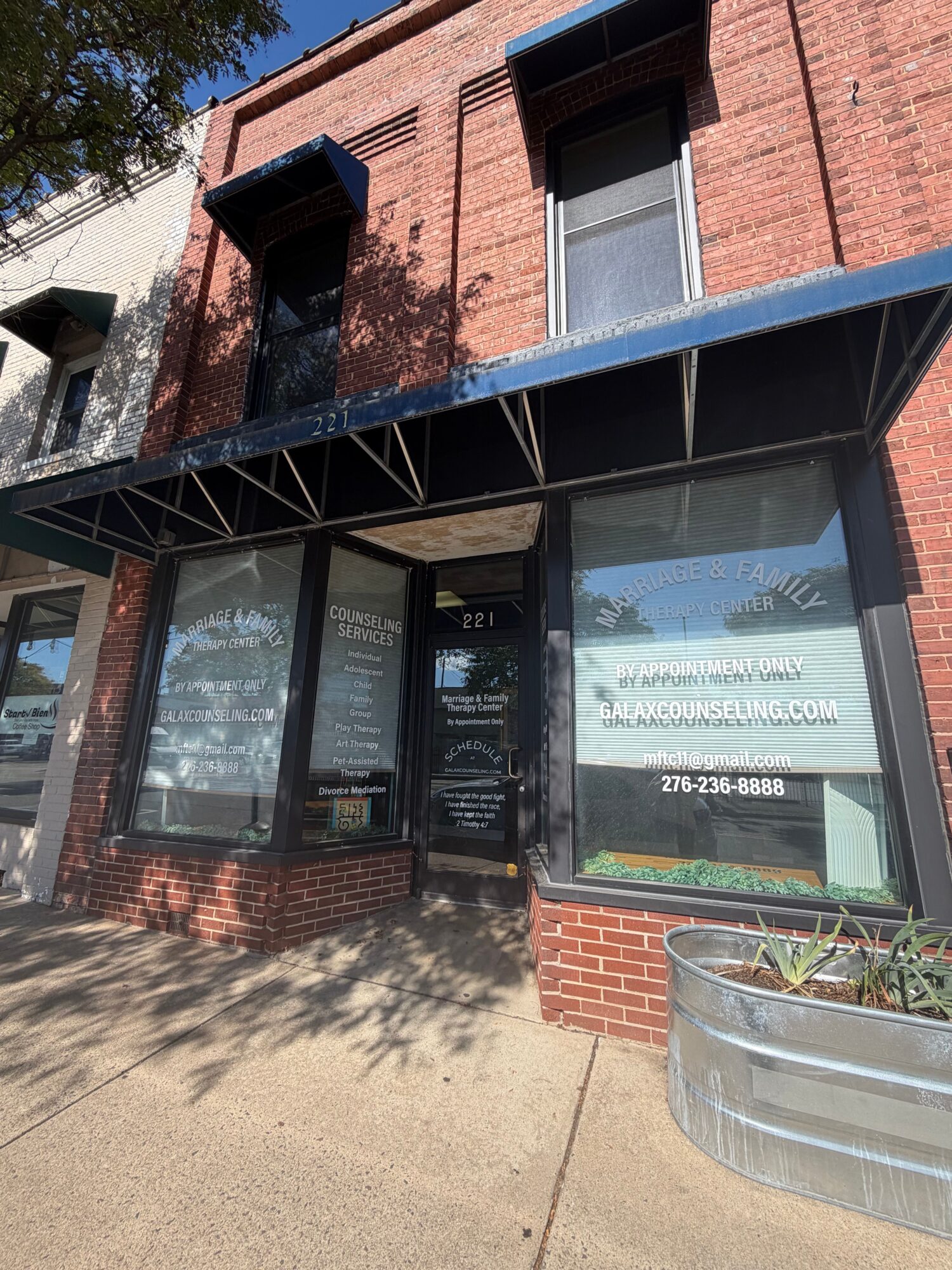 Storefront with glass door and window, brick exterior, sidewalk, and parked car, with shadows cast on the ground.