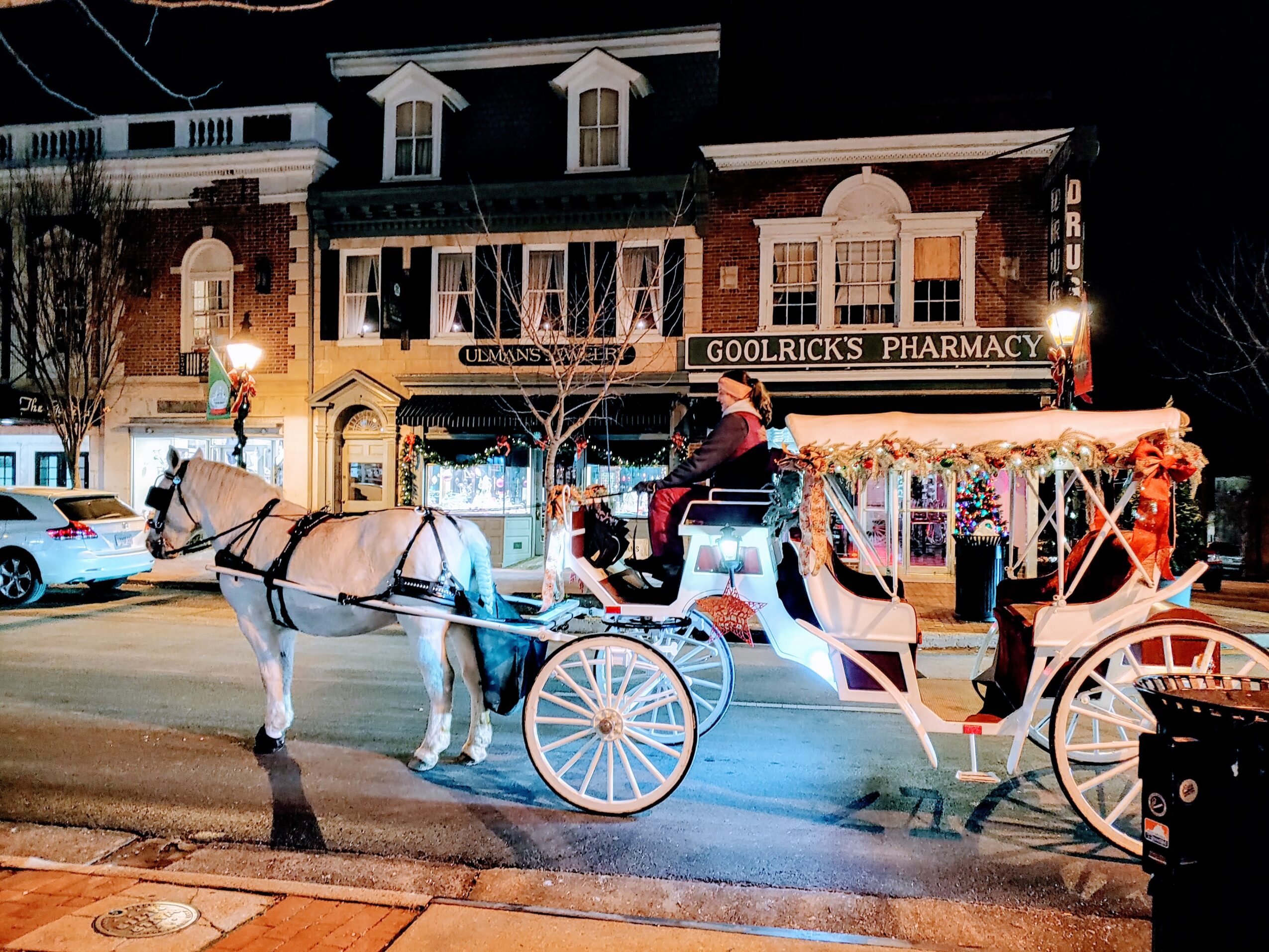 Horse-drawn carriage with illuminated decorations parked on a street at night, buildings and a pharmacy in the background.