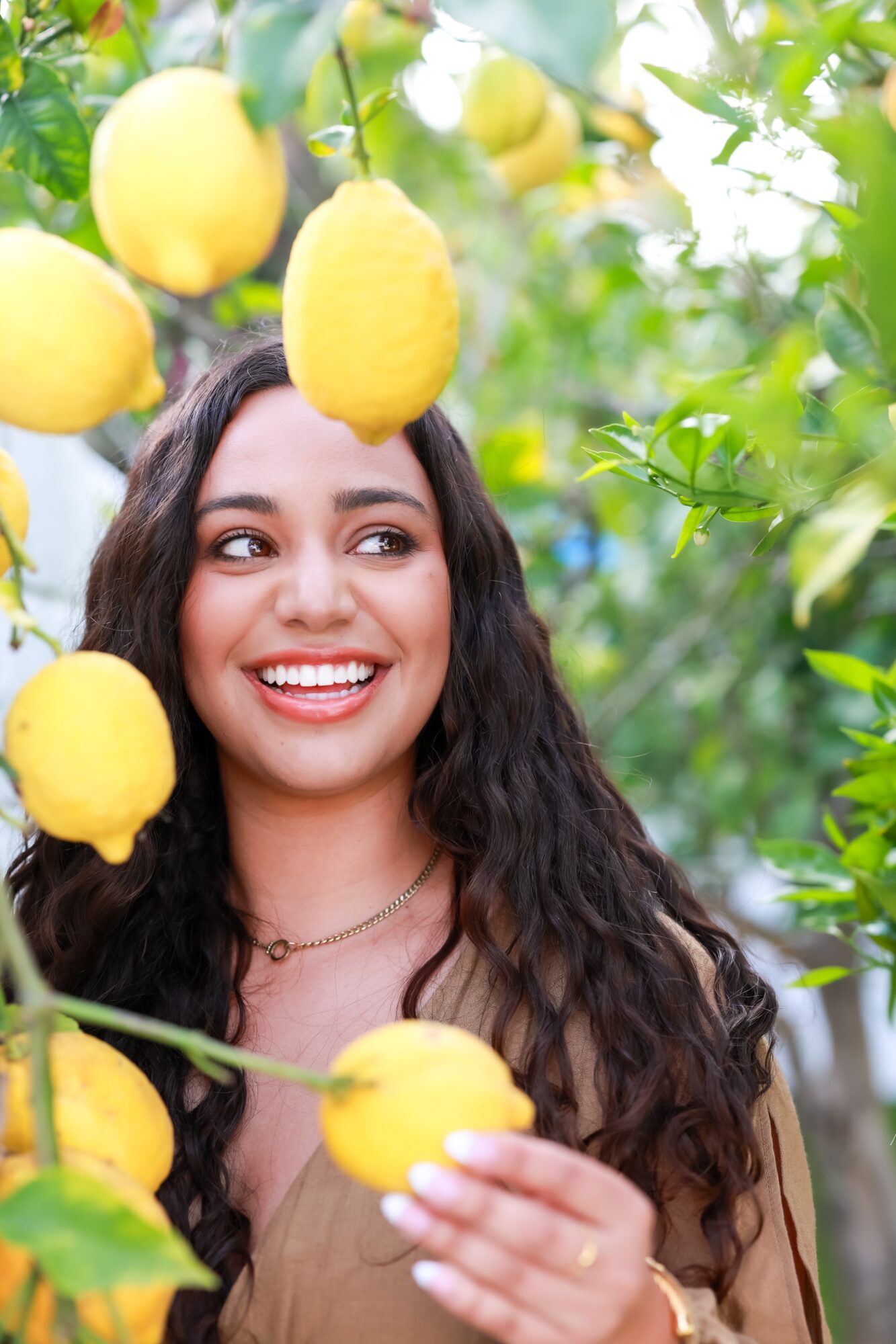 Smiling woman with long curly hair among lemon trees with ripe yellow lemons, surrounded by green leaves.