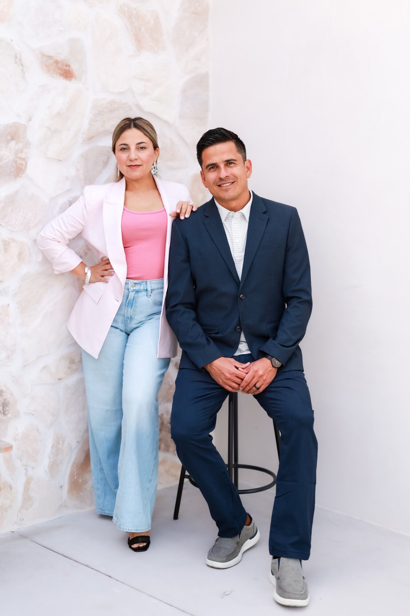 A woman and a man pose together indoors, woman standing with hand on hip, man sitting on a stool, both smiling.
