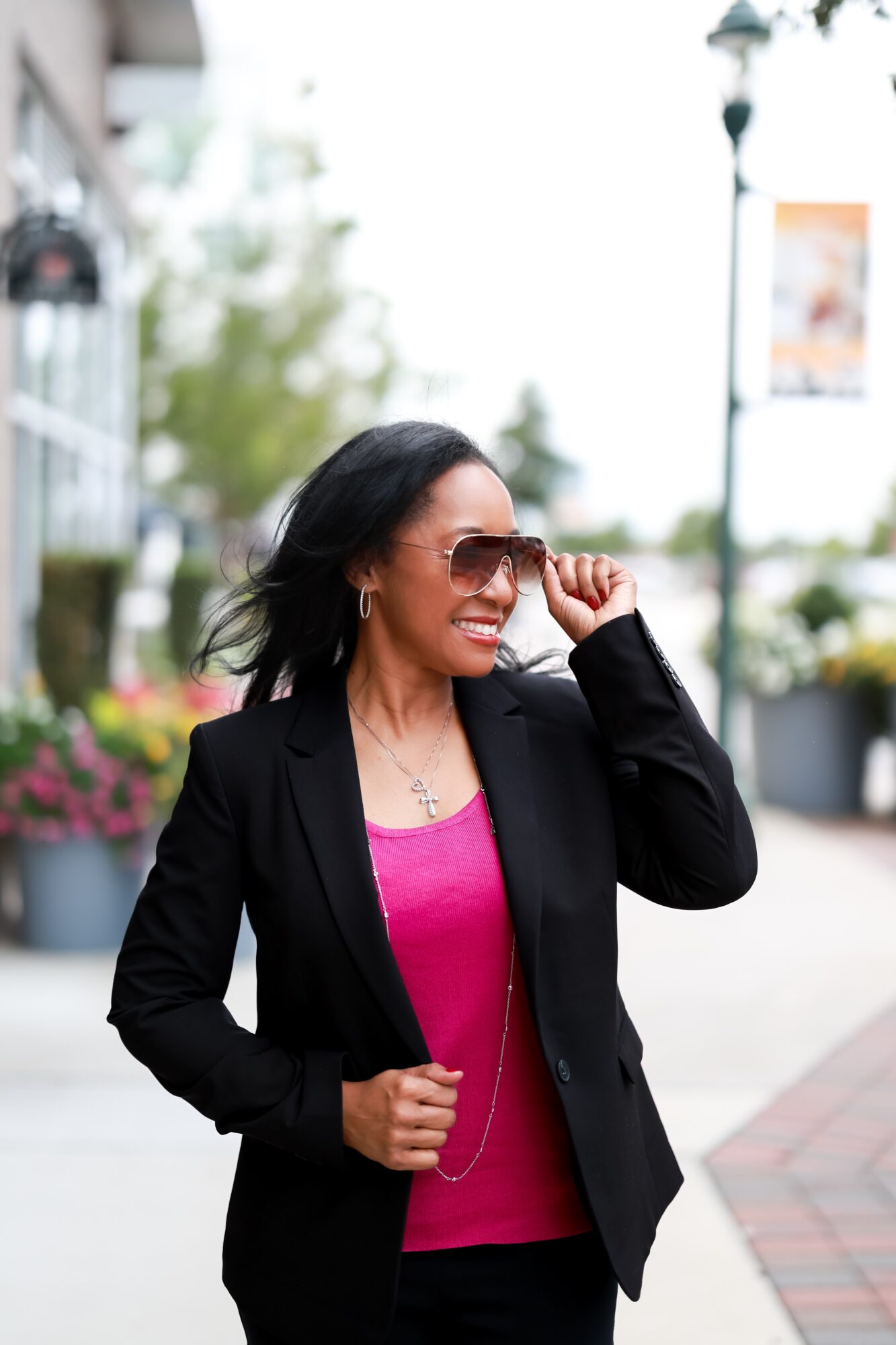 Woman with dark hair wearing sunglasses, black blazer, and pink top, smiling outdoors on a city sidewalk.
