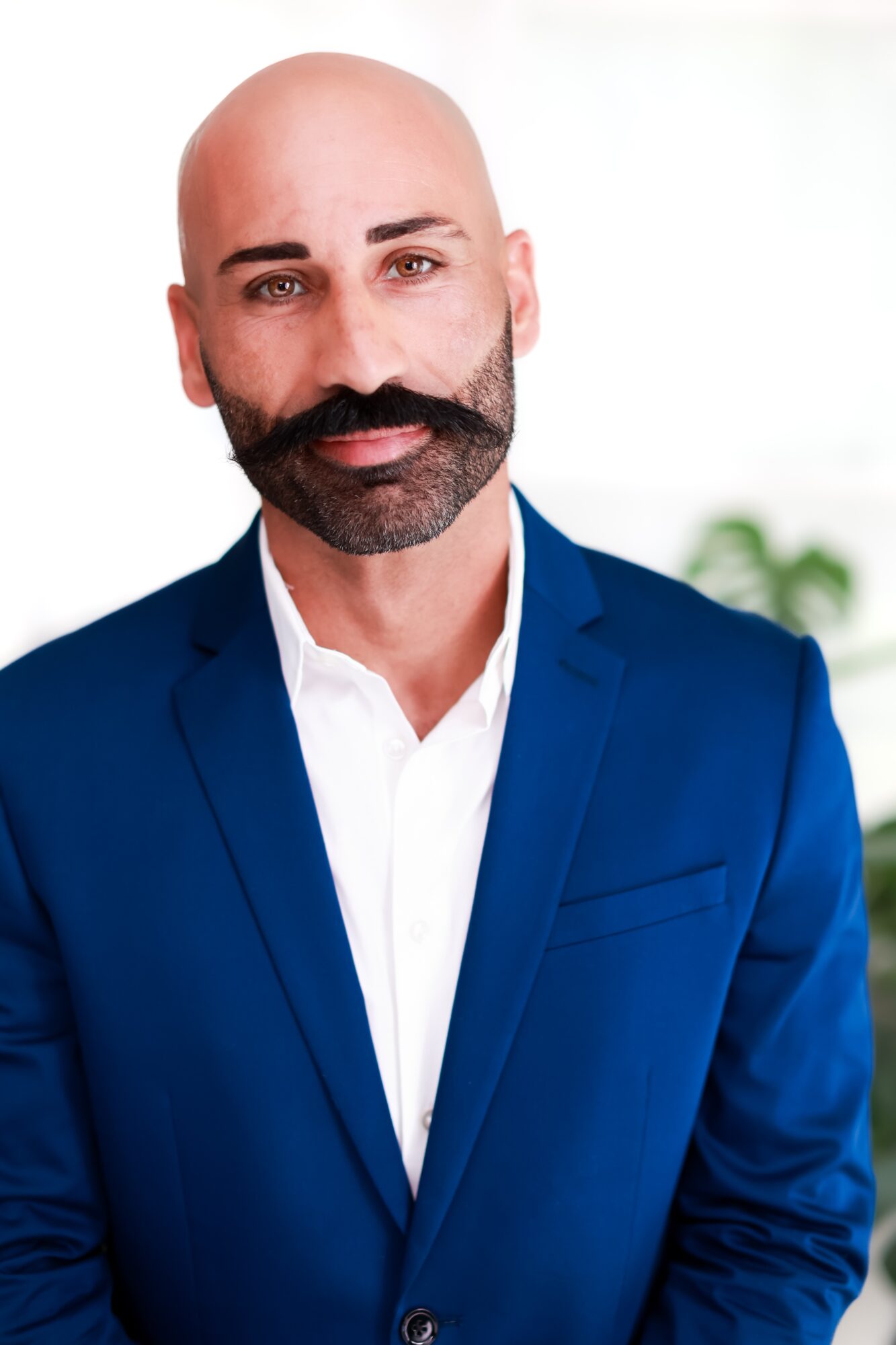 Bald man with a beard in a blue suit and white shirt, standing indoors with a blurred background.