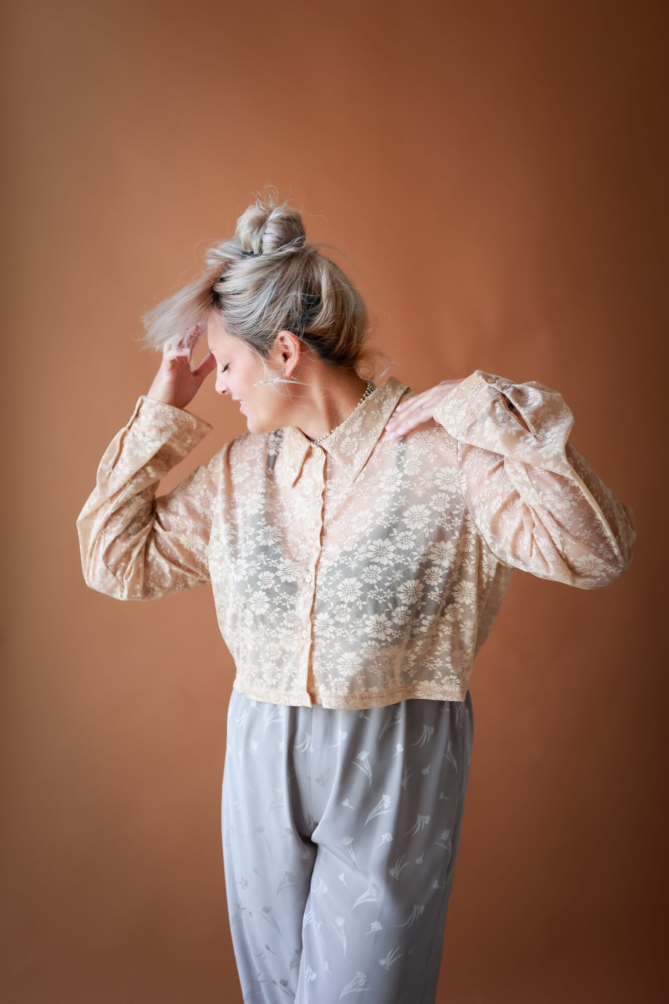 Woman with styled hair in a lace blouse and patterned pants, standing sideways with hand on her head, against a brown background.