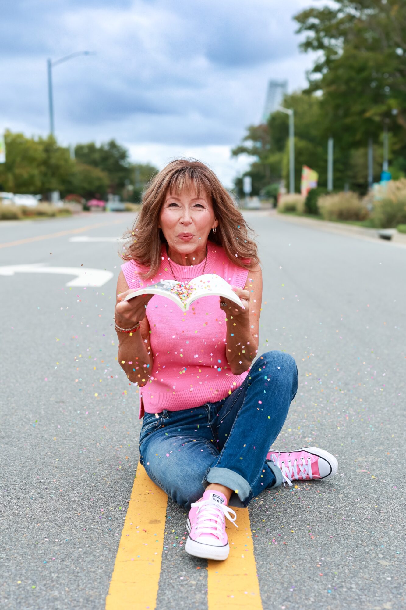 Woman sitting cross-legged on a street with trees and buildings in the background, holding a pink heart-shaped object, smiling.