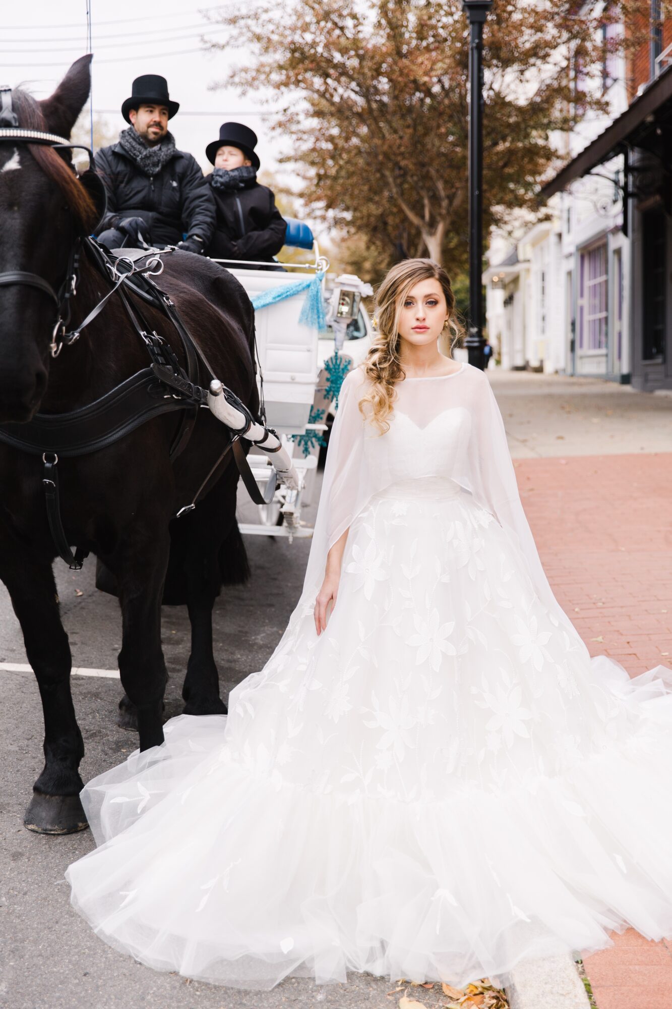 A bride in a white wedding gown and veil stands on a sidewalk, with a horse-drawn carriage and two men in the background.