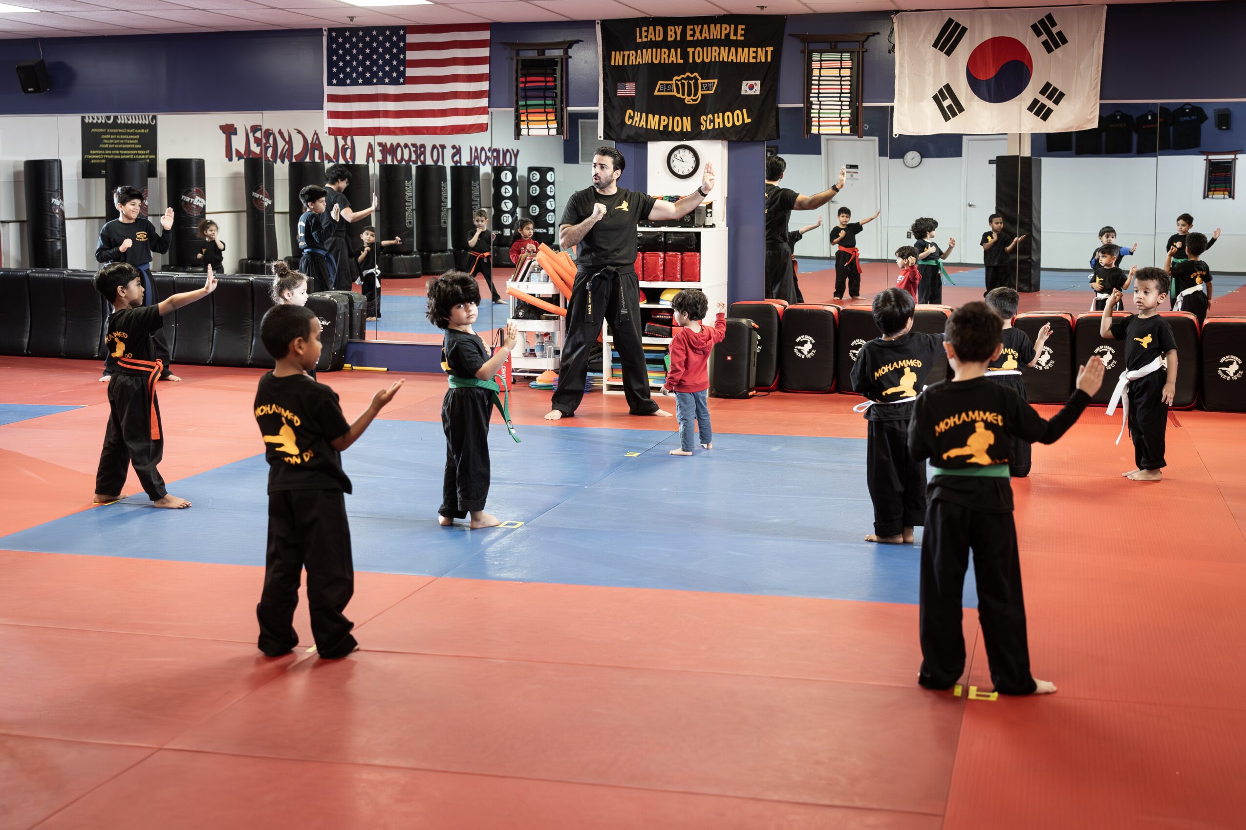 Children in martial arts uniforms practice in a dojo with instructors, mirrors, flags, and martial arts equipment visible.