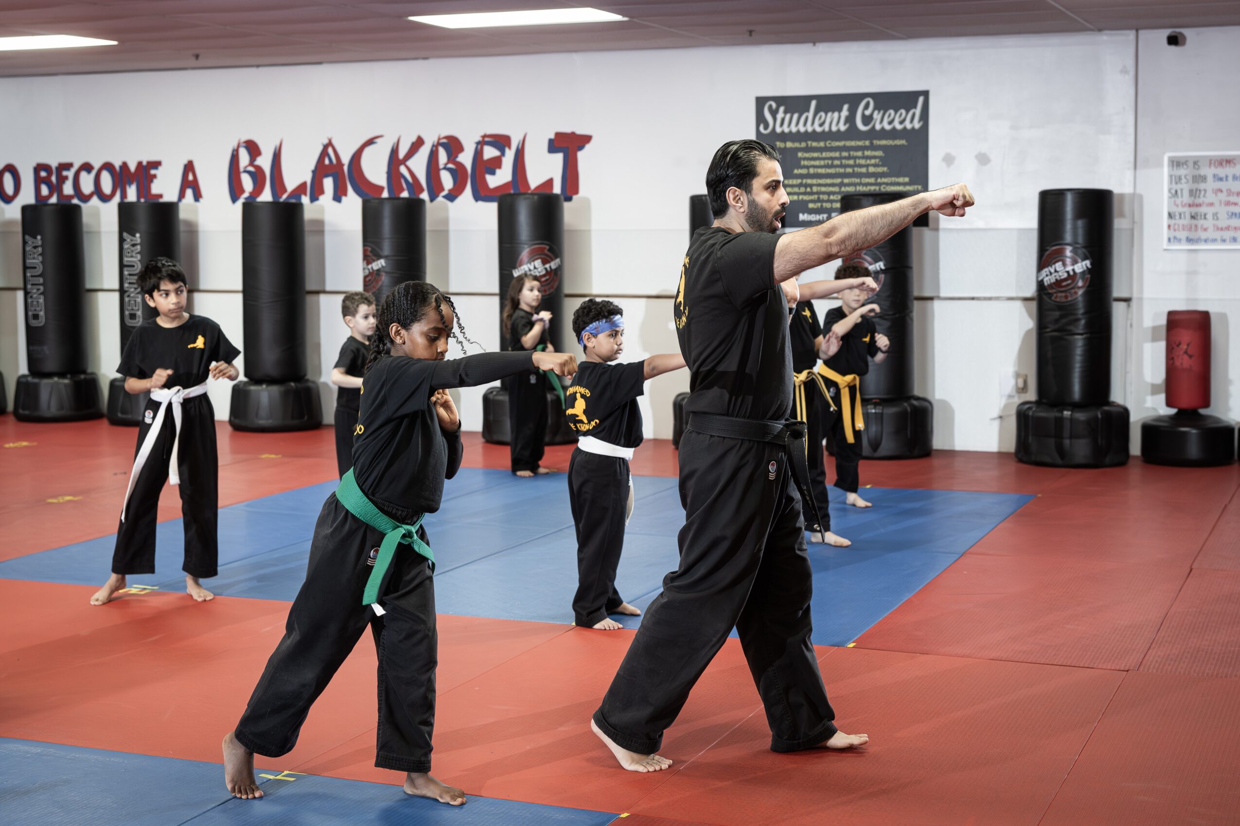 Instructor demonstrating martial arts techniques to children in a dojo with black punching bags and a sign reading 'How to Become a Blackbelt'.