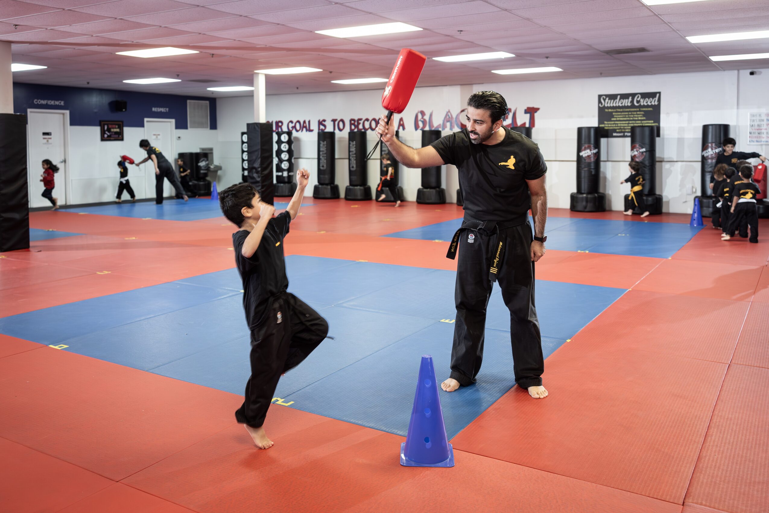 A martial arts instructor and a young student practicing in a gym with mats, cones, and other students in the background.