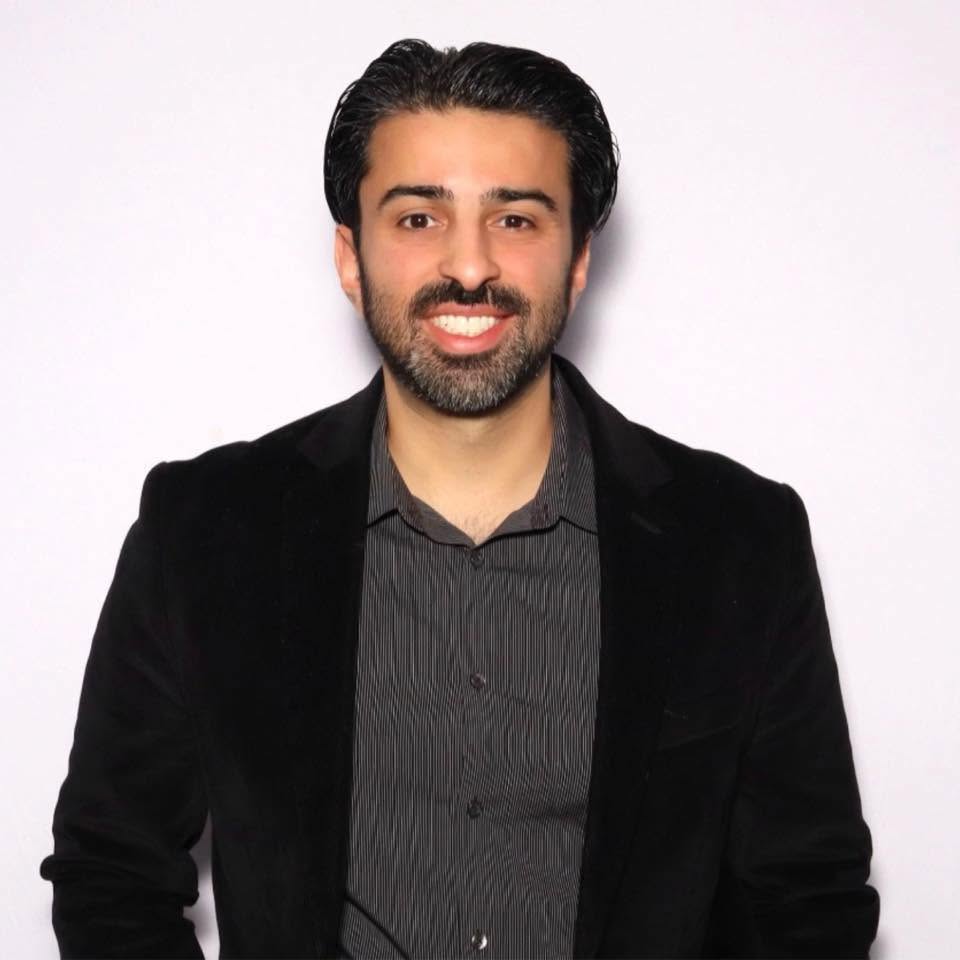 Man with dark hair and beard smiling, wearing a black blazer and dark shirt, against a white background.