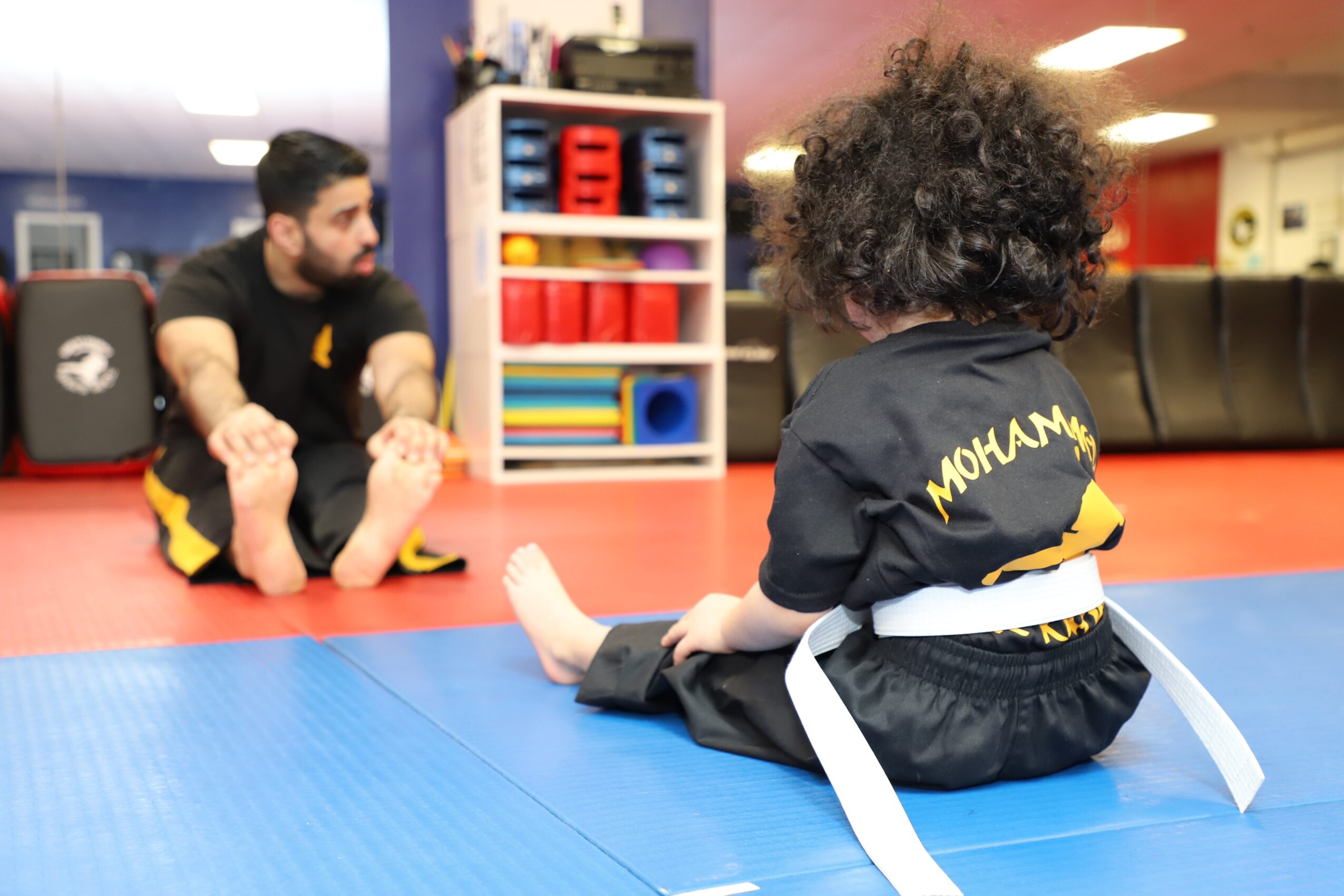 Child with curly hair sitting on a blue mat, wearing a black martial arts uniform with a white belt, facing an instructor in a martial arts studio.
