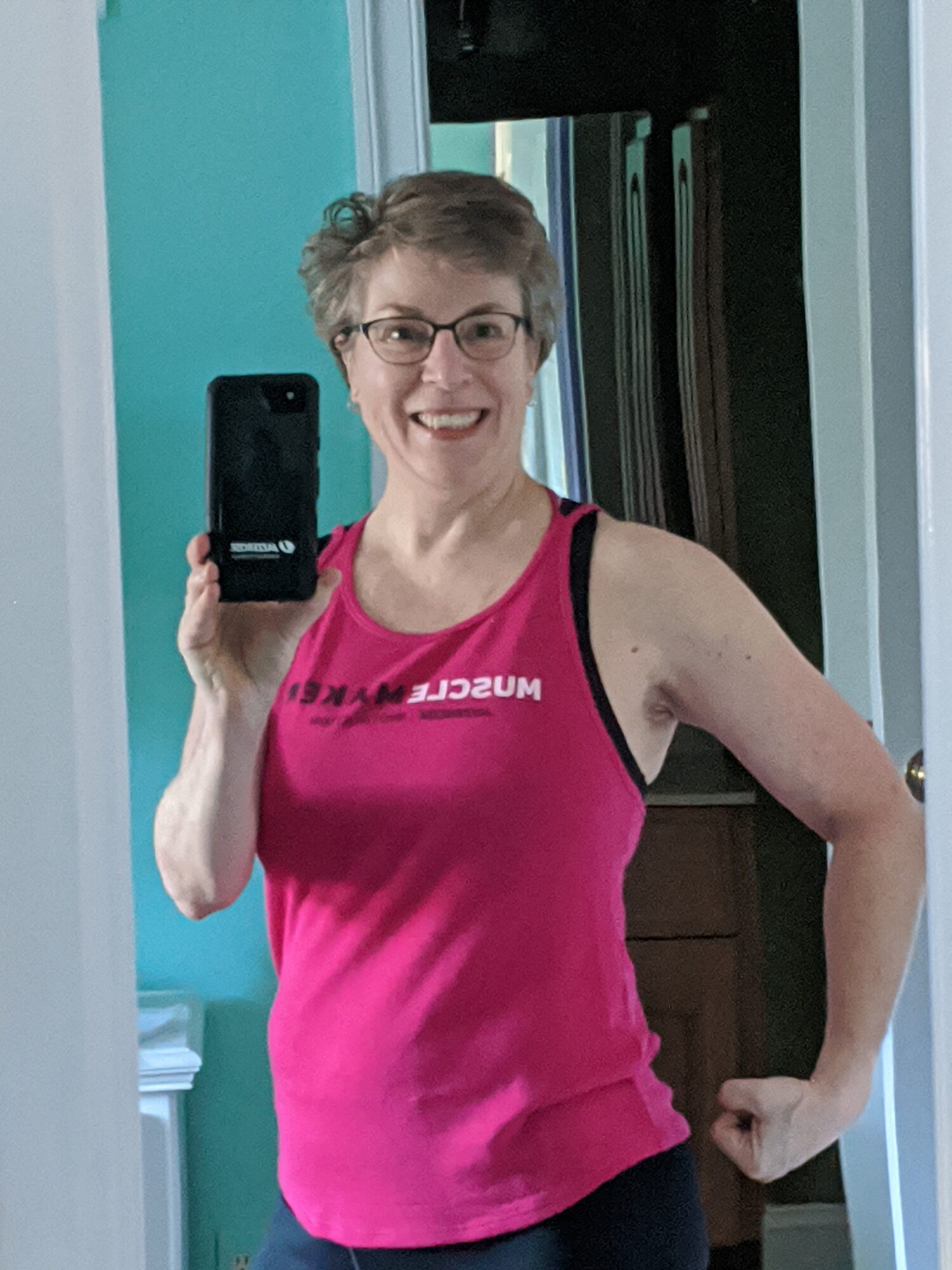 Smiling woman with short hair and glasses taking a mirror selfie, wearing a pink sleeveless top, in a room with teal walls.