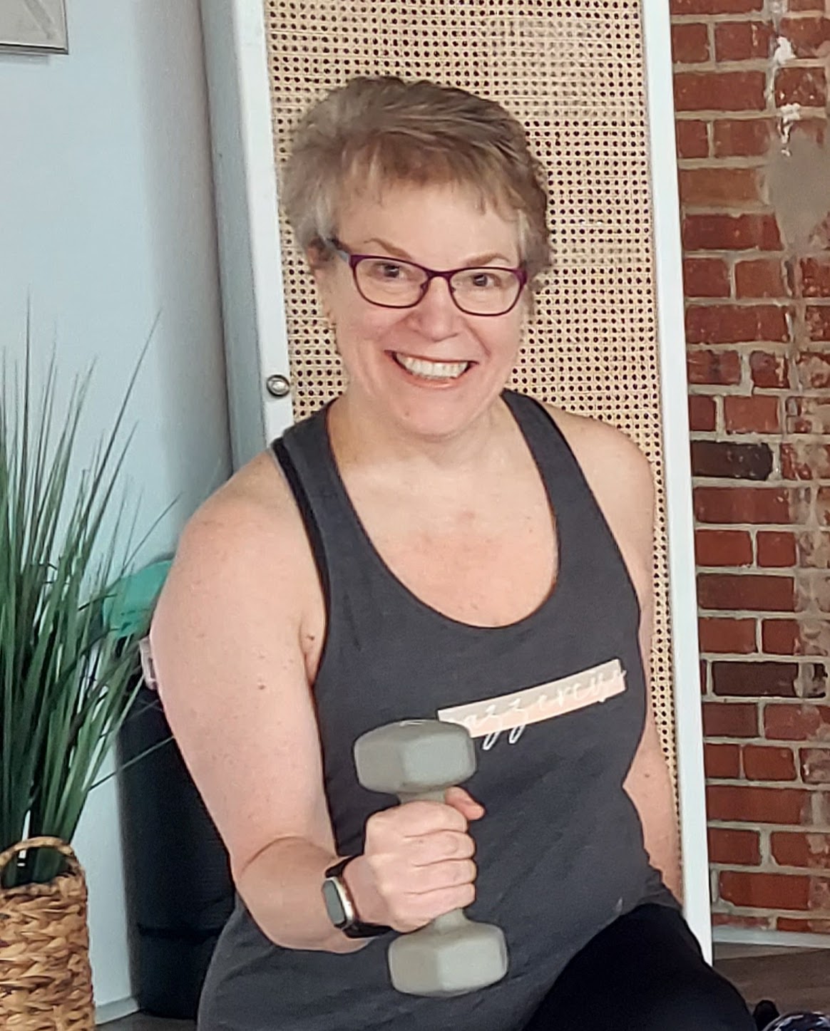 Smiling woman with short hair and glasses holding a gray dumbbell indoors, wearing a dark tank top.
