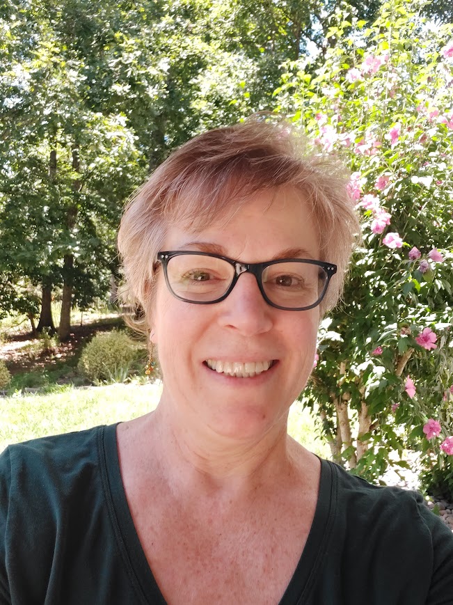 Woman with short hair and glasses smiling outdoors with trees and pink flowers in background.