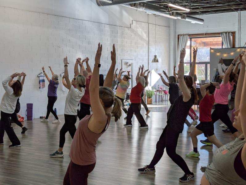 Group of people participating in a fitness class with arms raised, in a spacious indoor studio with large windows.