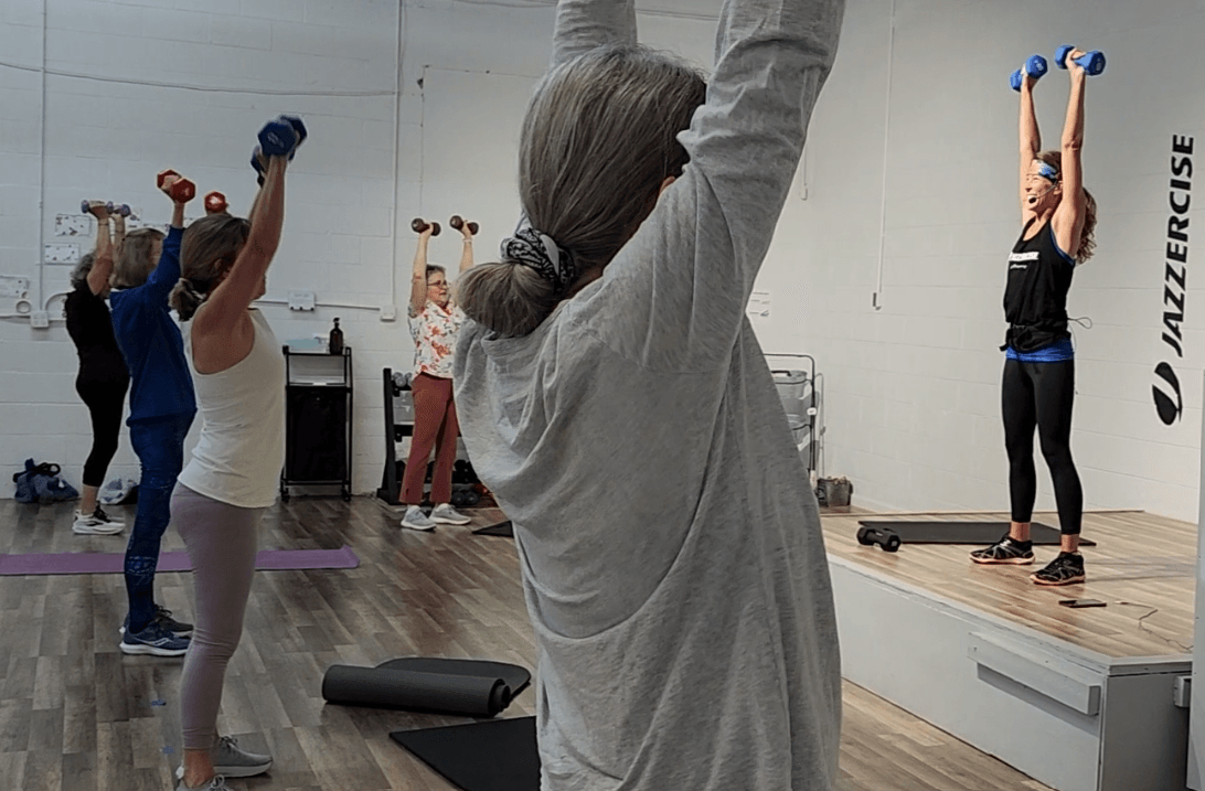 Group of people participating in a fitness class, lifting dumbbells overhead in a studio with a trainer on a platform.