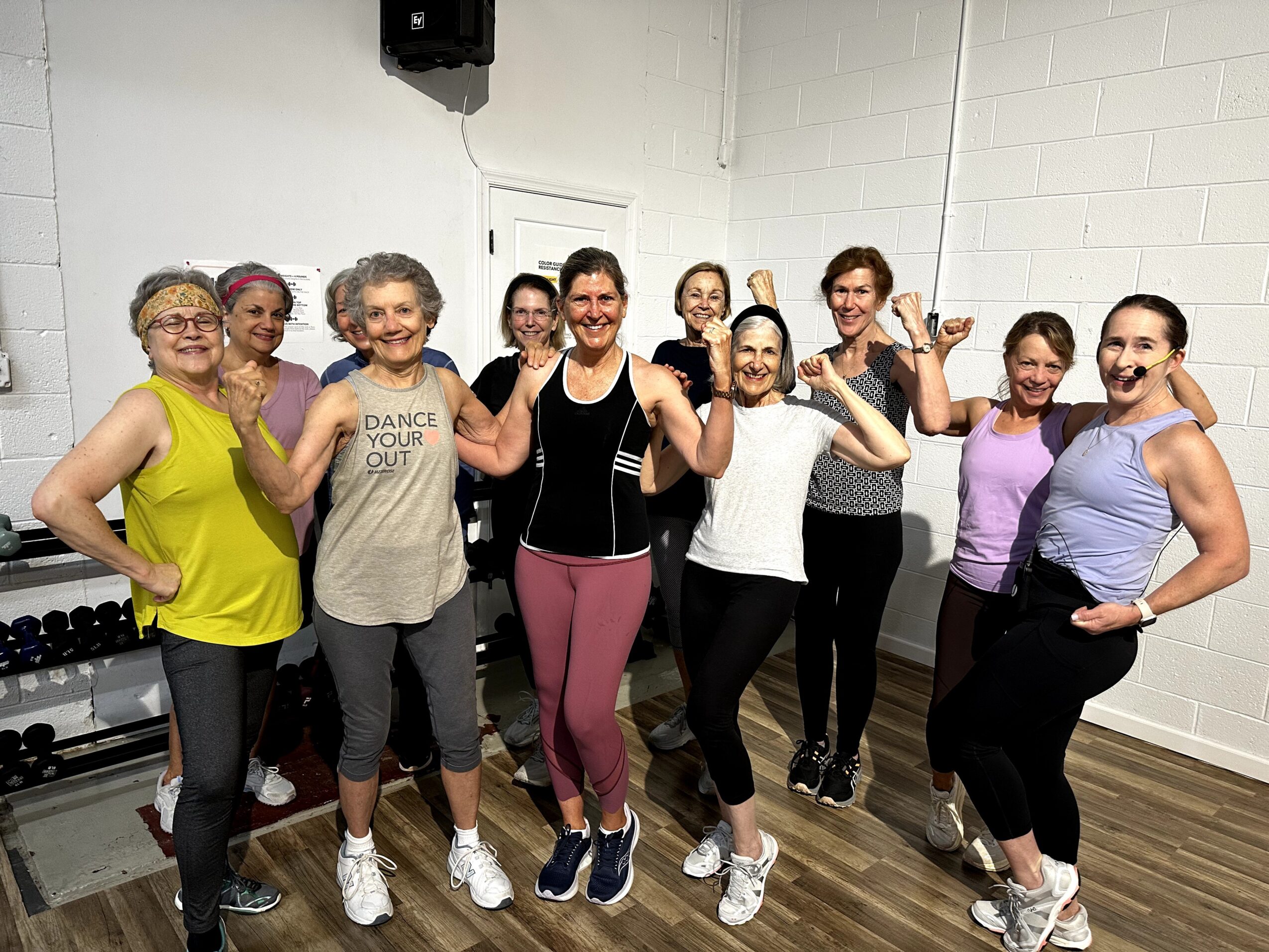 Group of nine women in workout clothes flexing their arms in a fitness class, smiling and showing strength.