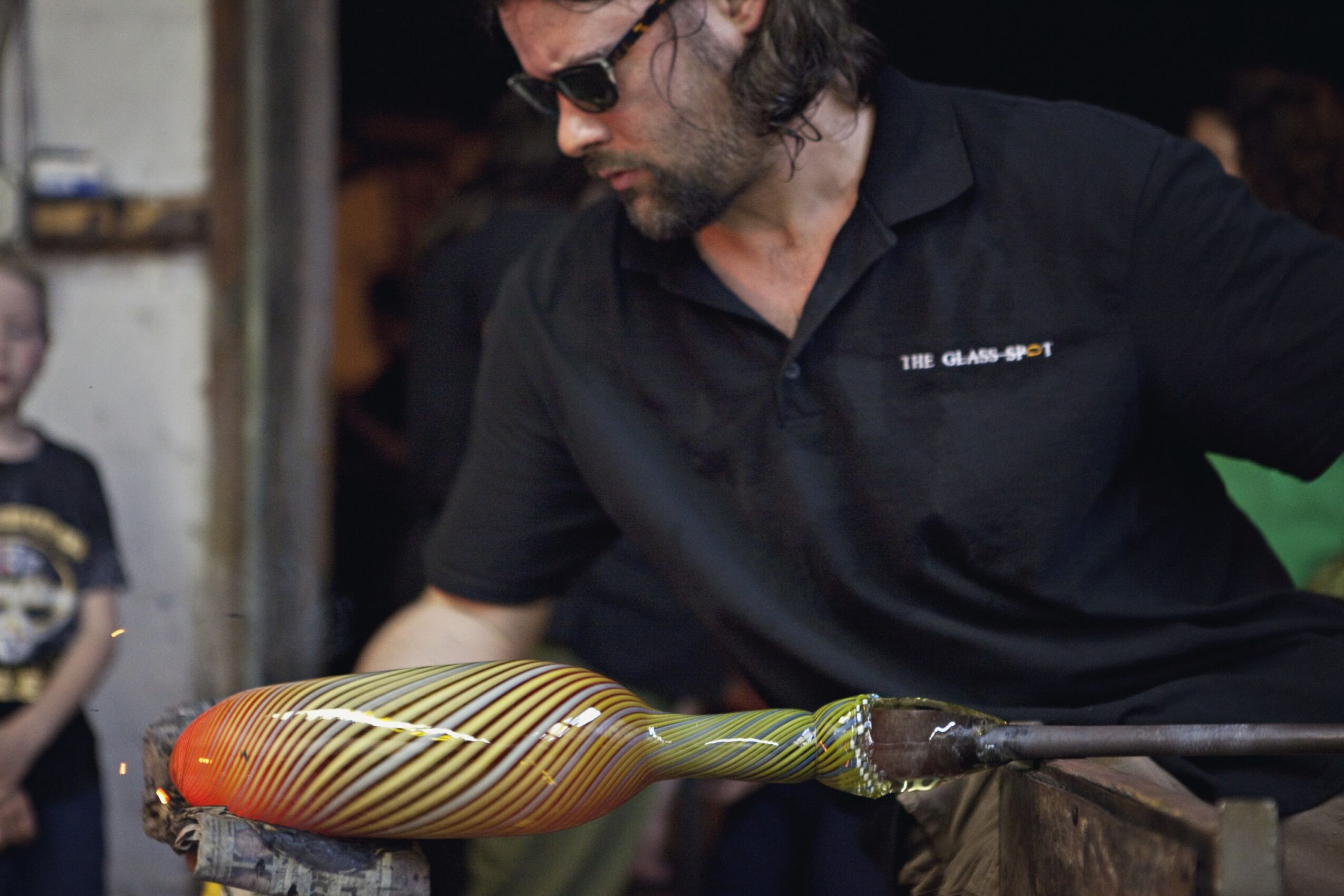Man with sunglasses working on a glassblowing project, holding a colorful glass object, in a workshop setting.