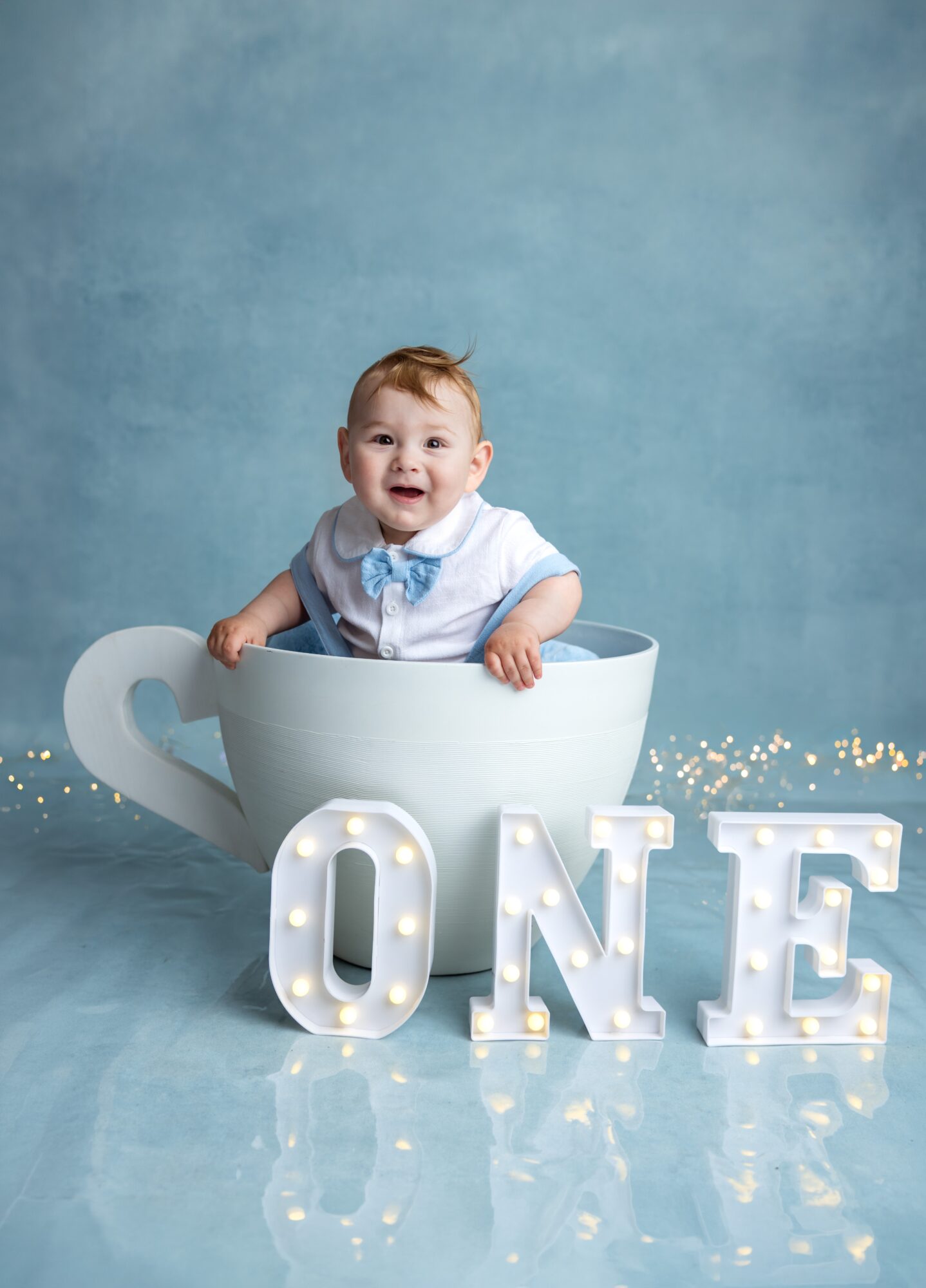 Baby in a large teacup with illuminated letters spelling 'ONE' in front, celebrating first birthday, against a blue background.