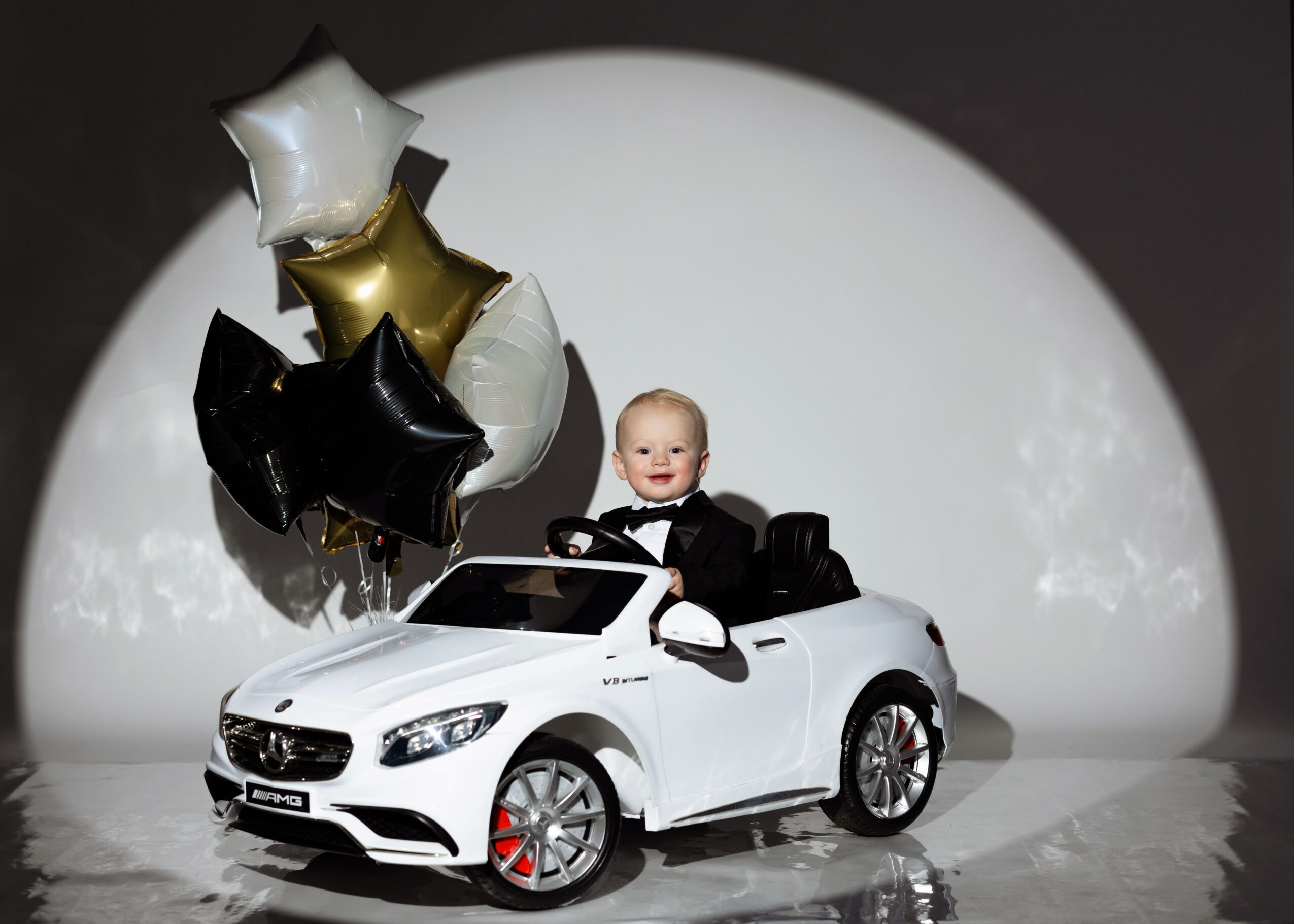 Child sitting in a white toy car with balloons behind, on a stage with a white circular backdrop.