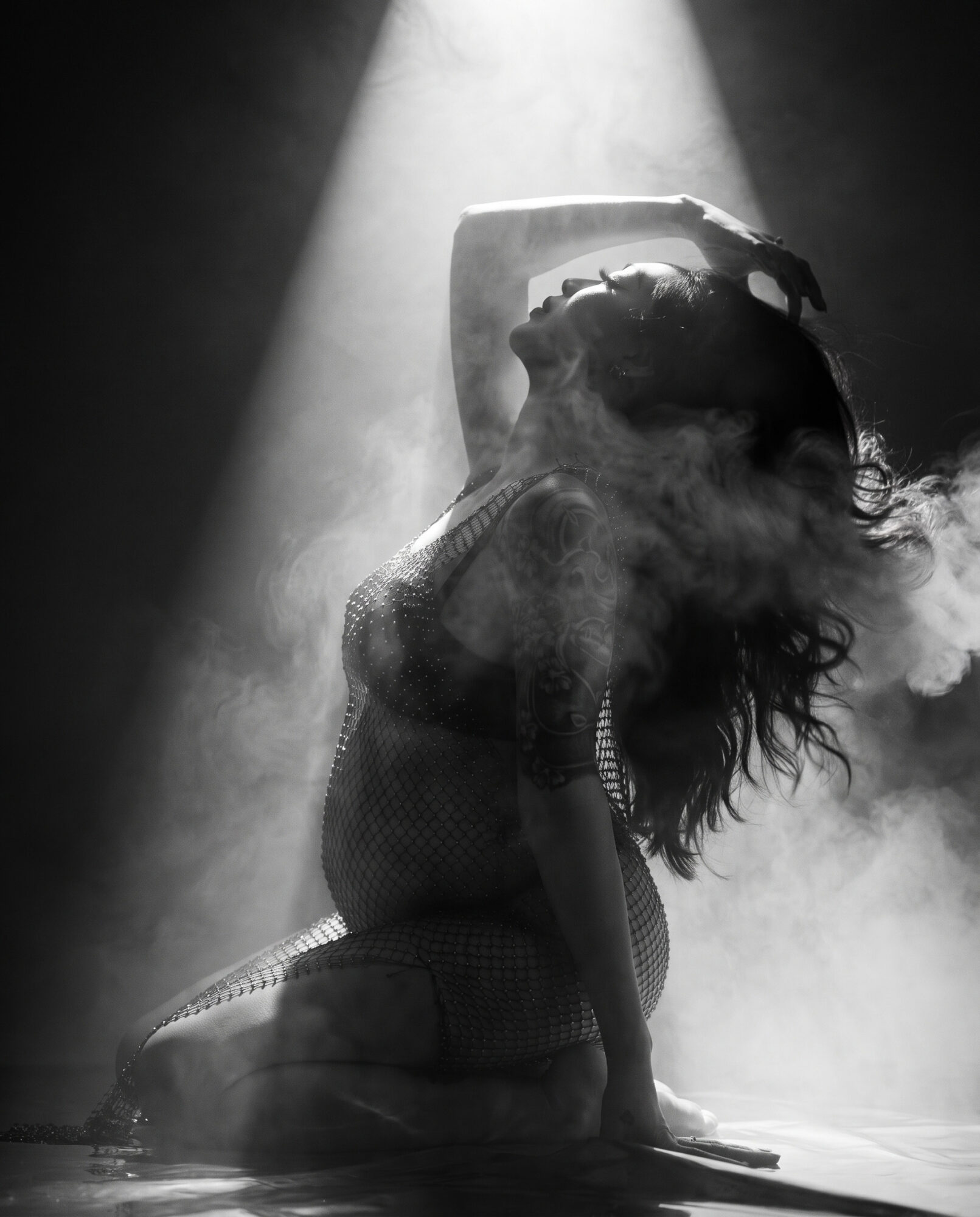 Person with long hair in a fishnet outfit, head tilted back, illuminated by a spotlight with smoke around.