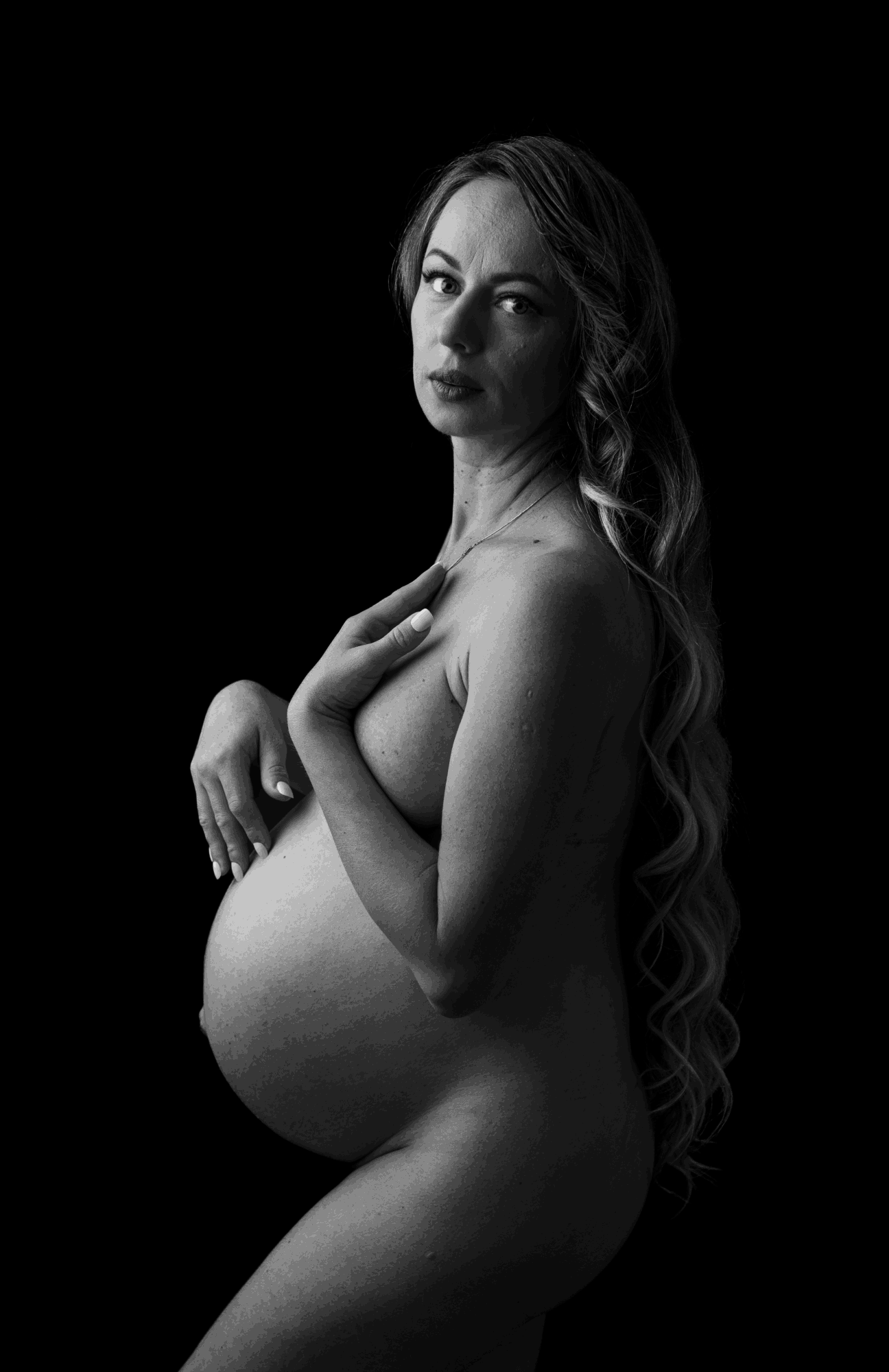 Pregnant woman with long hair, looking at the camera, hand on her chest, in black and white.