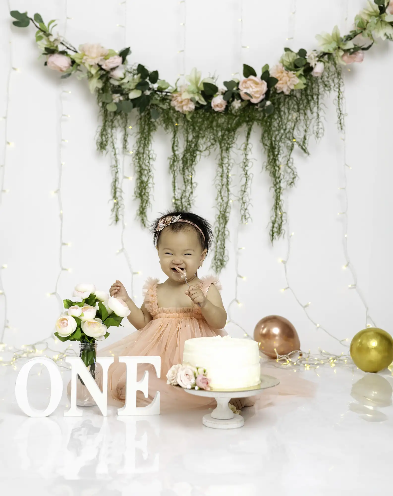 Child in pink dress celebrating first birthday with cake, flowers, and balloons, decorated with floral garland and hanging greenery.