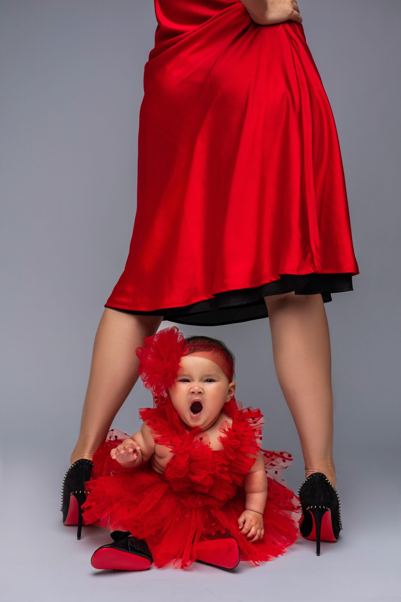 Child in red dress and headband sitting on the floor, with a woman standing behind her, legs visible.