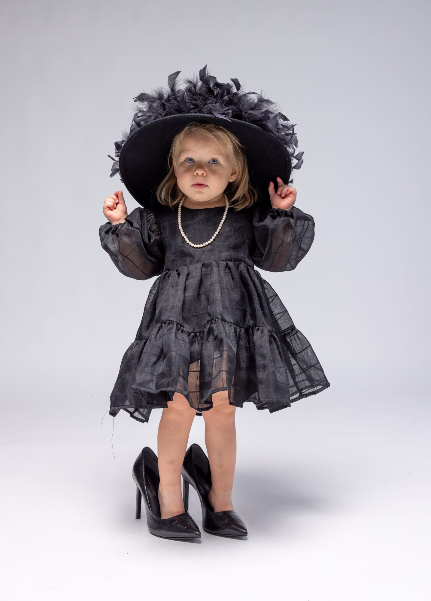 Young girl in a black dress and large hat with feathers, wearing high heels, standing against a plain background.