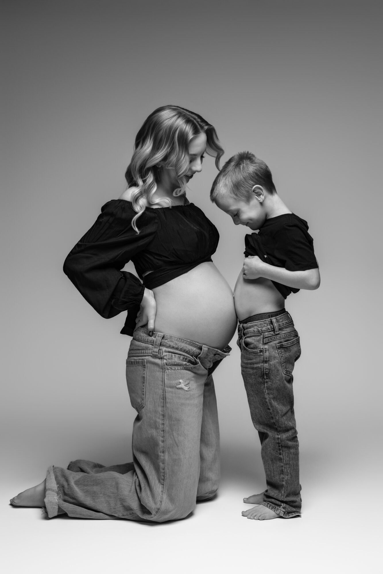 Pregnant woman and young boy looking at her belly, both wearing black shirts and jeans, standing against a plain background.