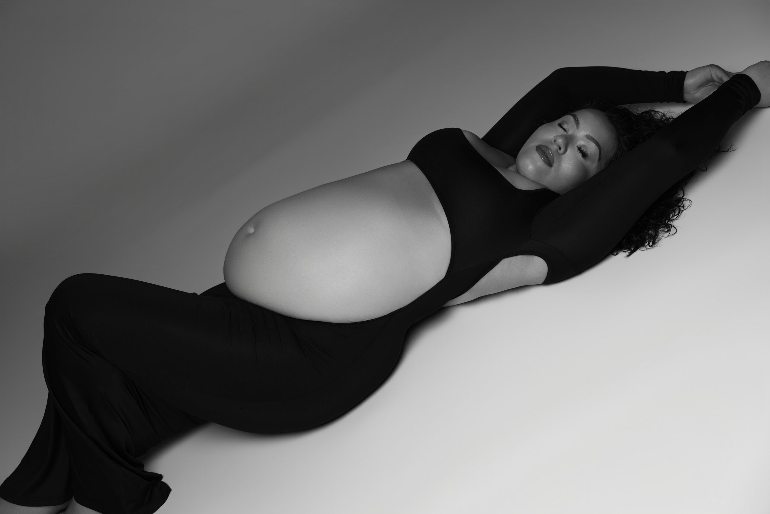 Woman lying on her back with arms raised above her head, wearing a black outfit, in black and white photo.