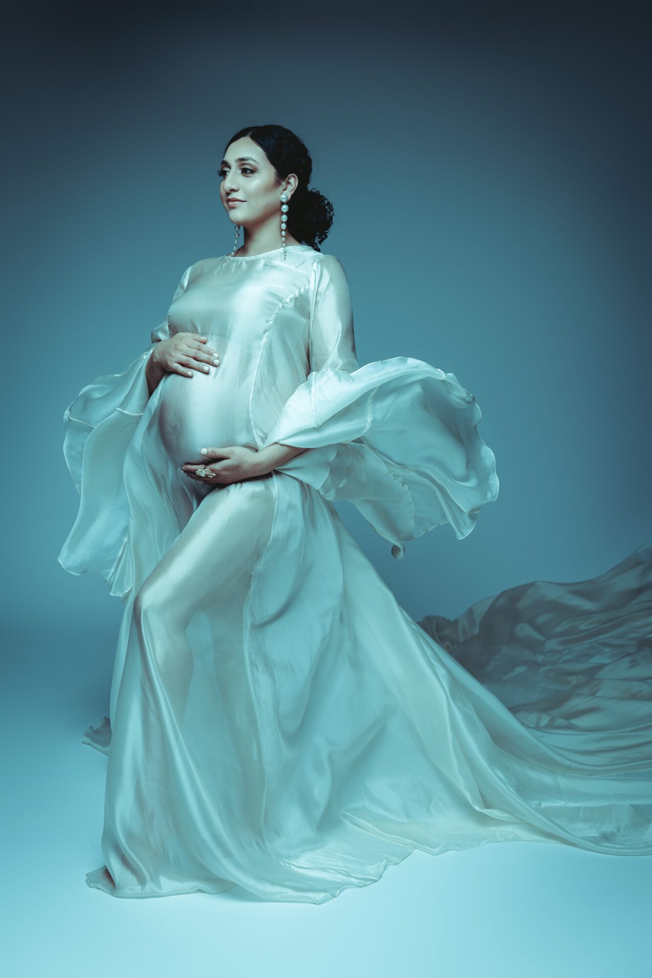 Pregnant woman in flowing white dress holding her belly, standing against a dark background with fabric on the ground.