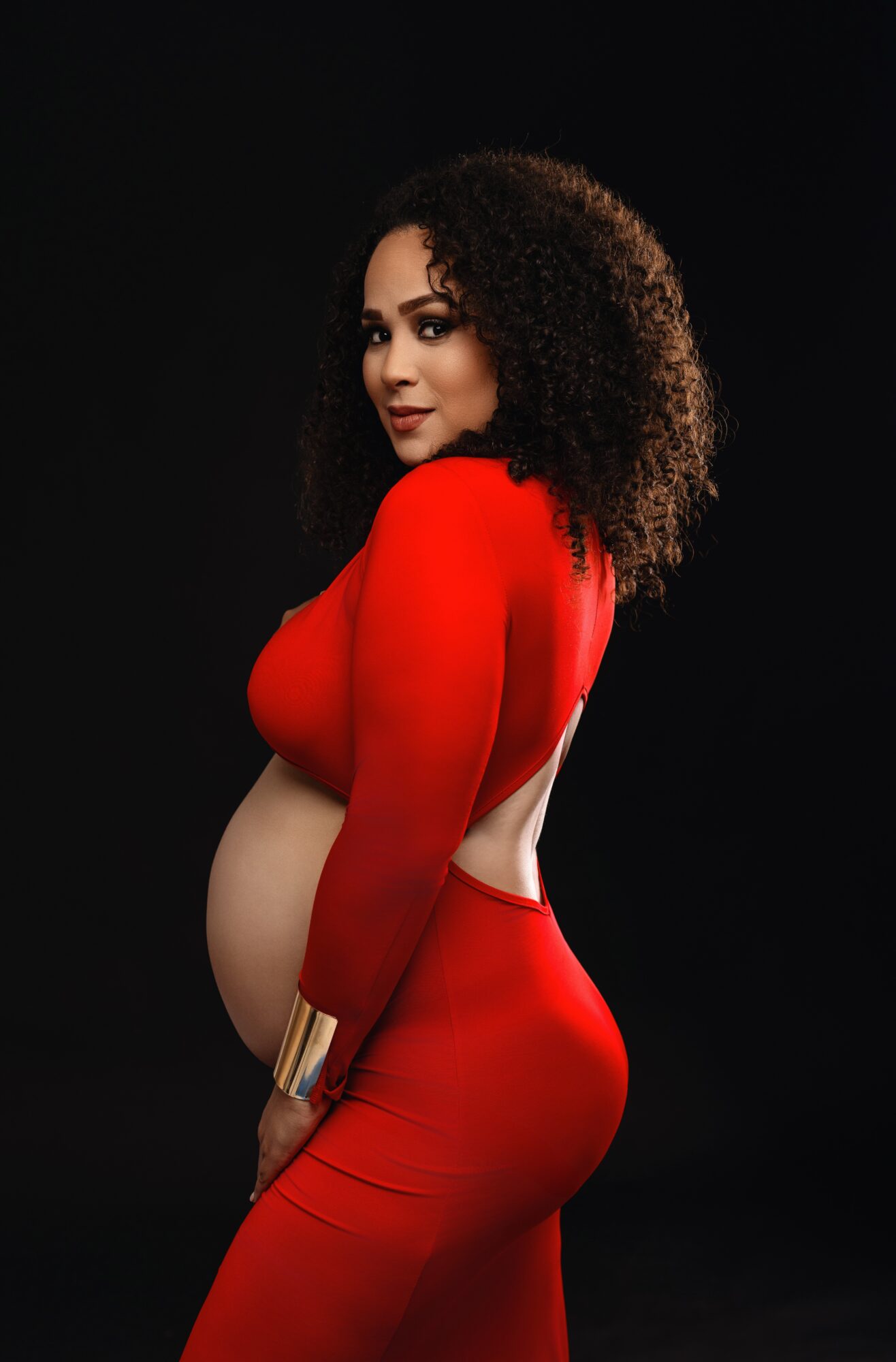 Woman with curly hair in a red dress with open back, standing sideways against a black background.