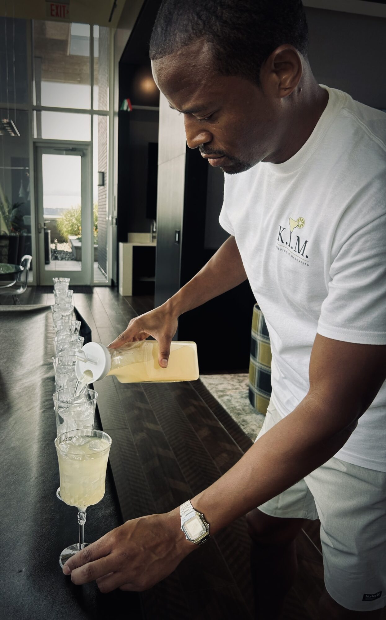Person pouring a drink into a glass at a bar counter, with a window in the background.