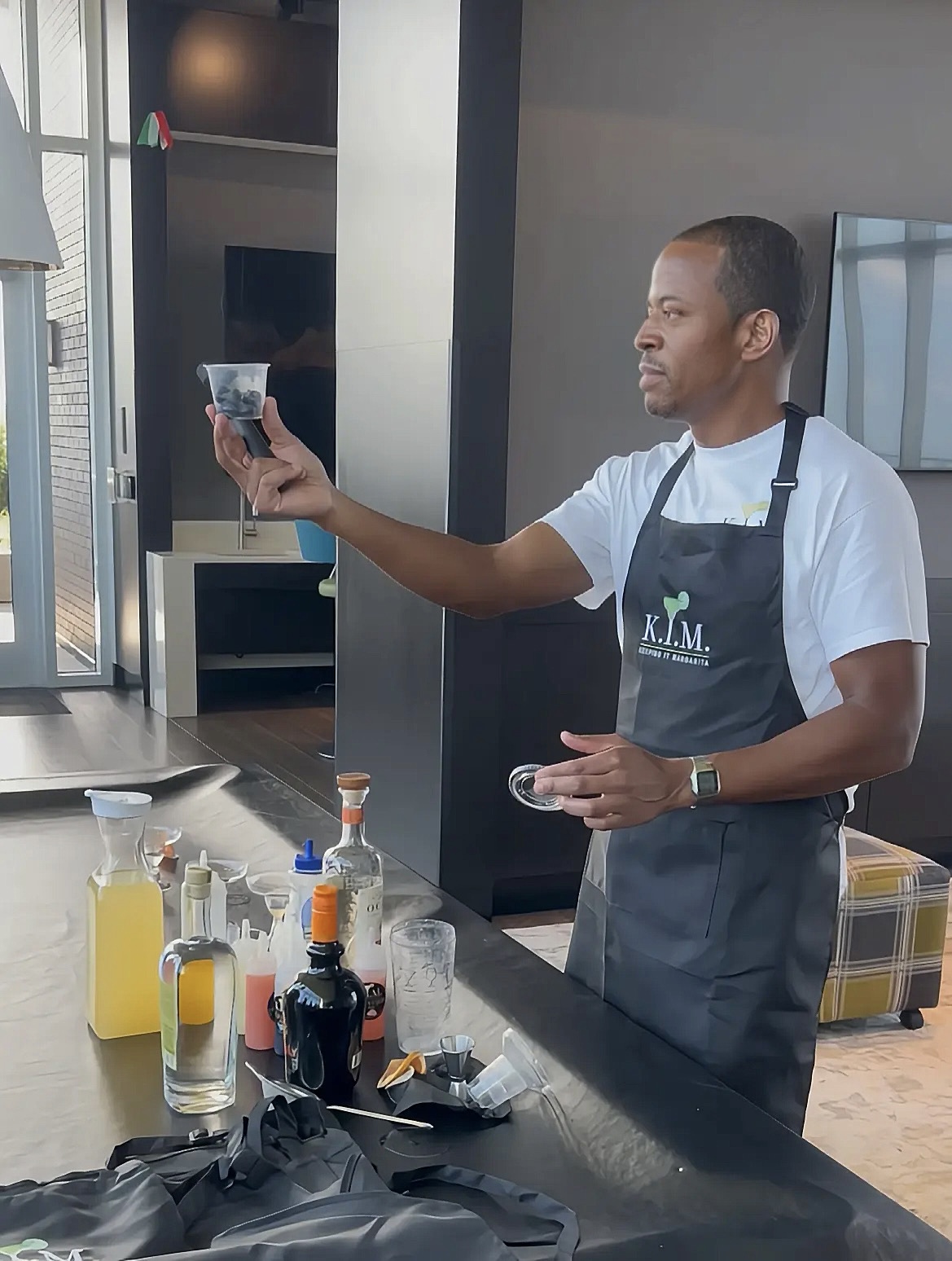 Man in apron taking a selfie in a modern kitchen with bottles and utensils on the counter.