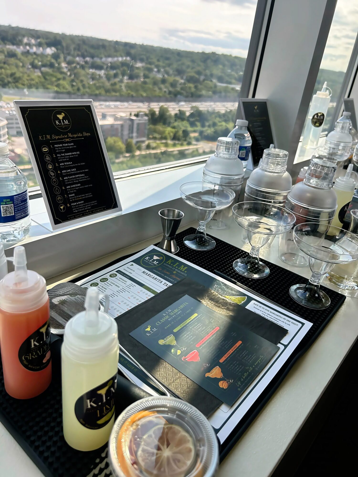 Barista station with bottles, glasses, and a tablet on a counter near a window with city view.