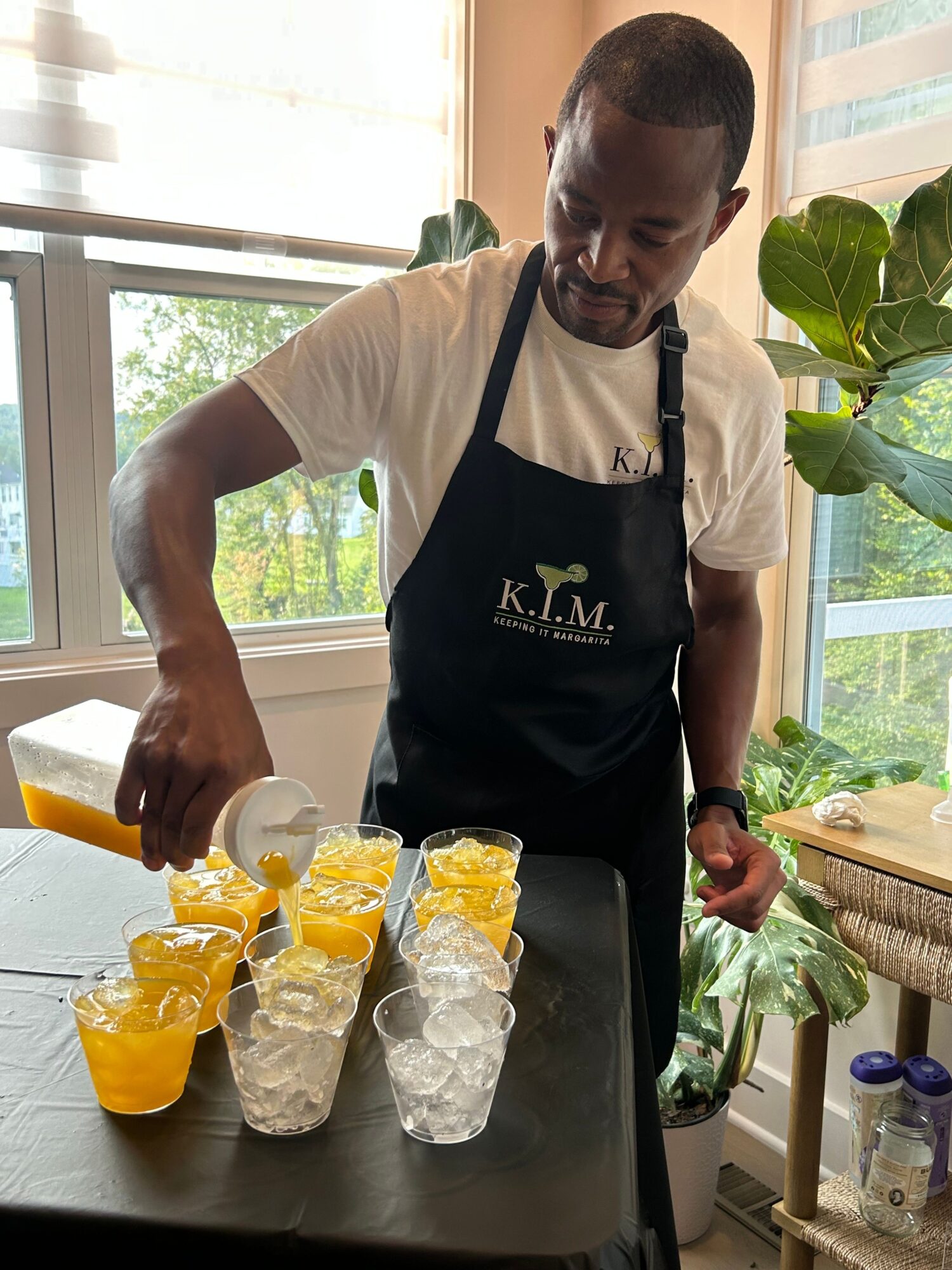 Person pouring a yellow drink into a glass, with bottles of water and lemon slices on the table, in a bright room.