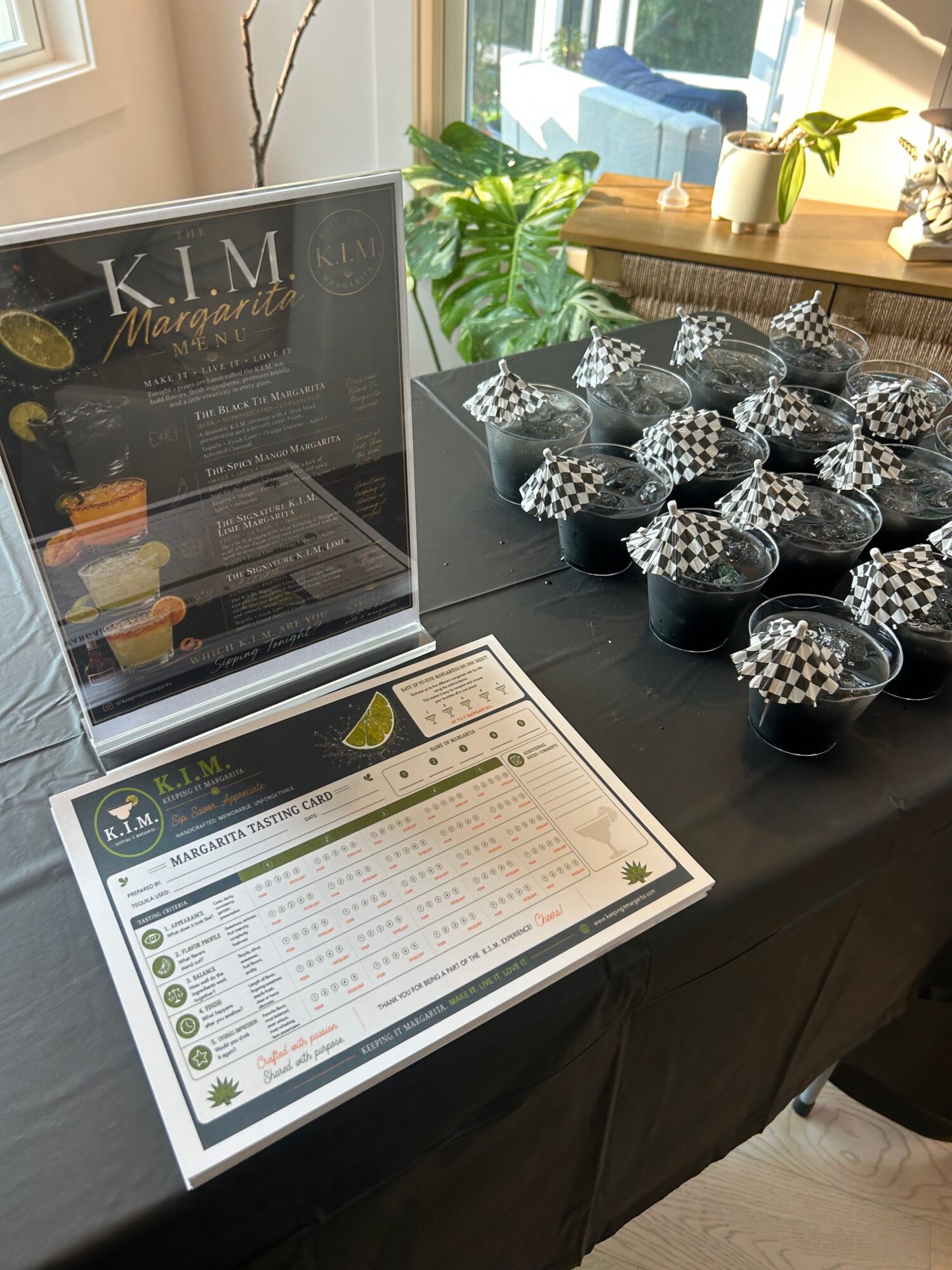 Display of multiple small checkered paper cups on a black table with a menu and a plant nearby.