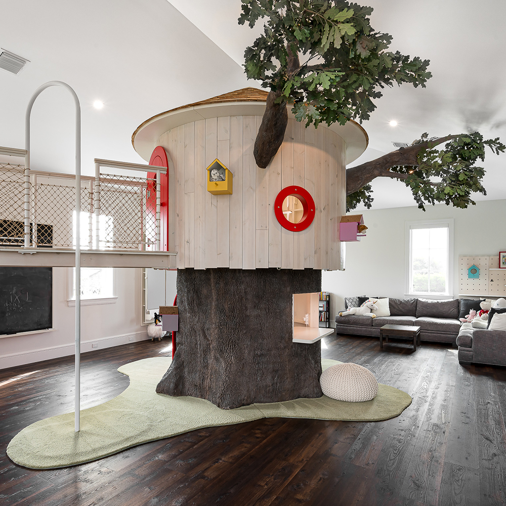Indoor playhouse shaped like a tree with a rounded wooden structure, windows, and a tree branch extending from the top, in a spacious room with dark wood flooring and a gray sofa.