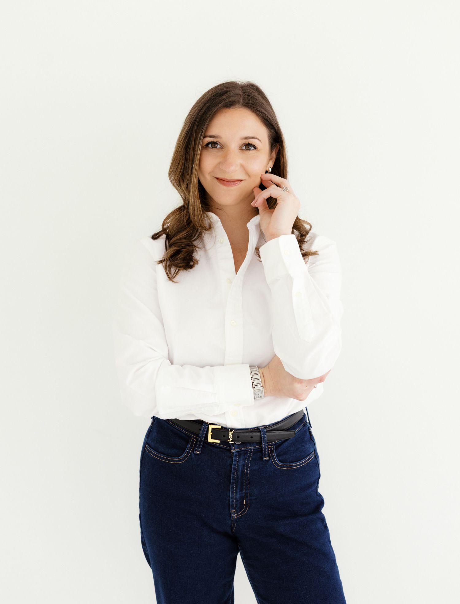Woman with brown hair in a white shirt and dark jeans, smiling and talking on a phone, standing against a white background.