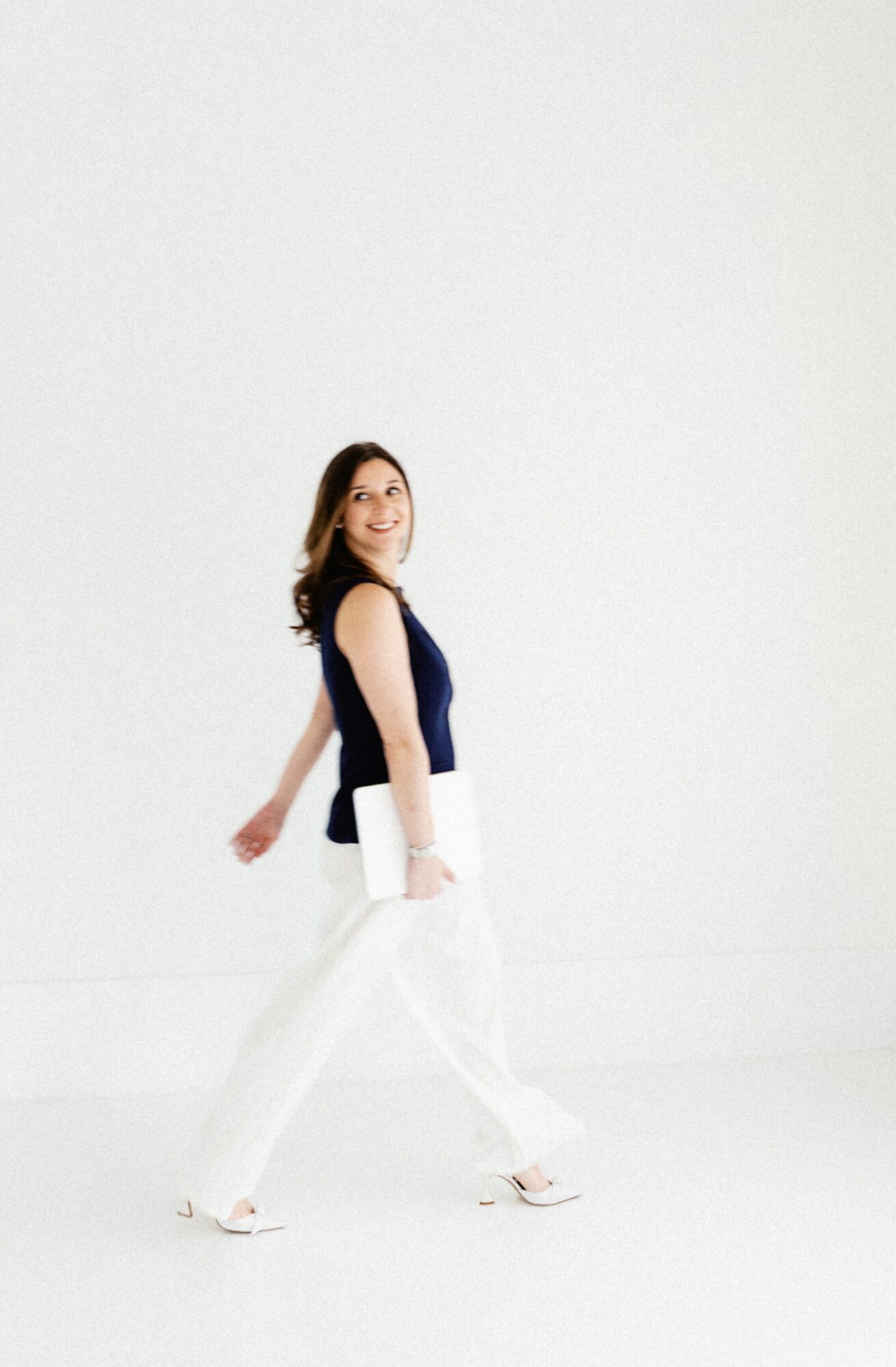 Woman walking with a smile, wearing a dark top and white pants, against a plain white background.