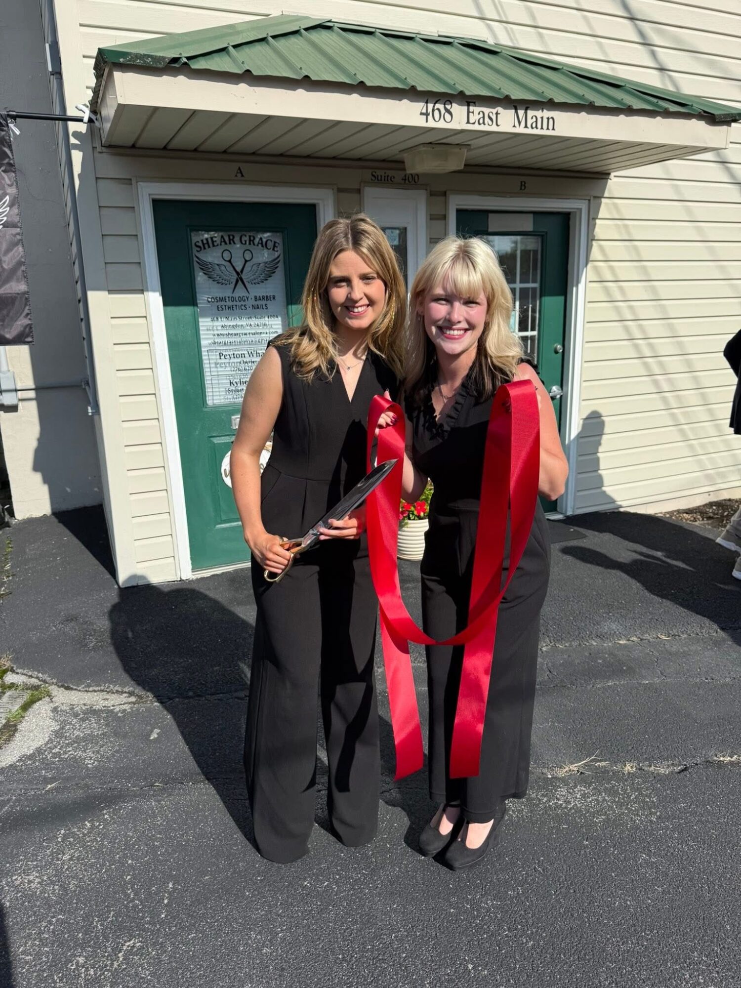 Two women standing outside a building, smiling, holding a large red ribbon and scissors, celebrating an event.