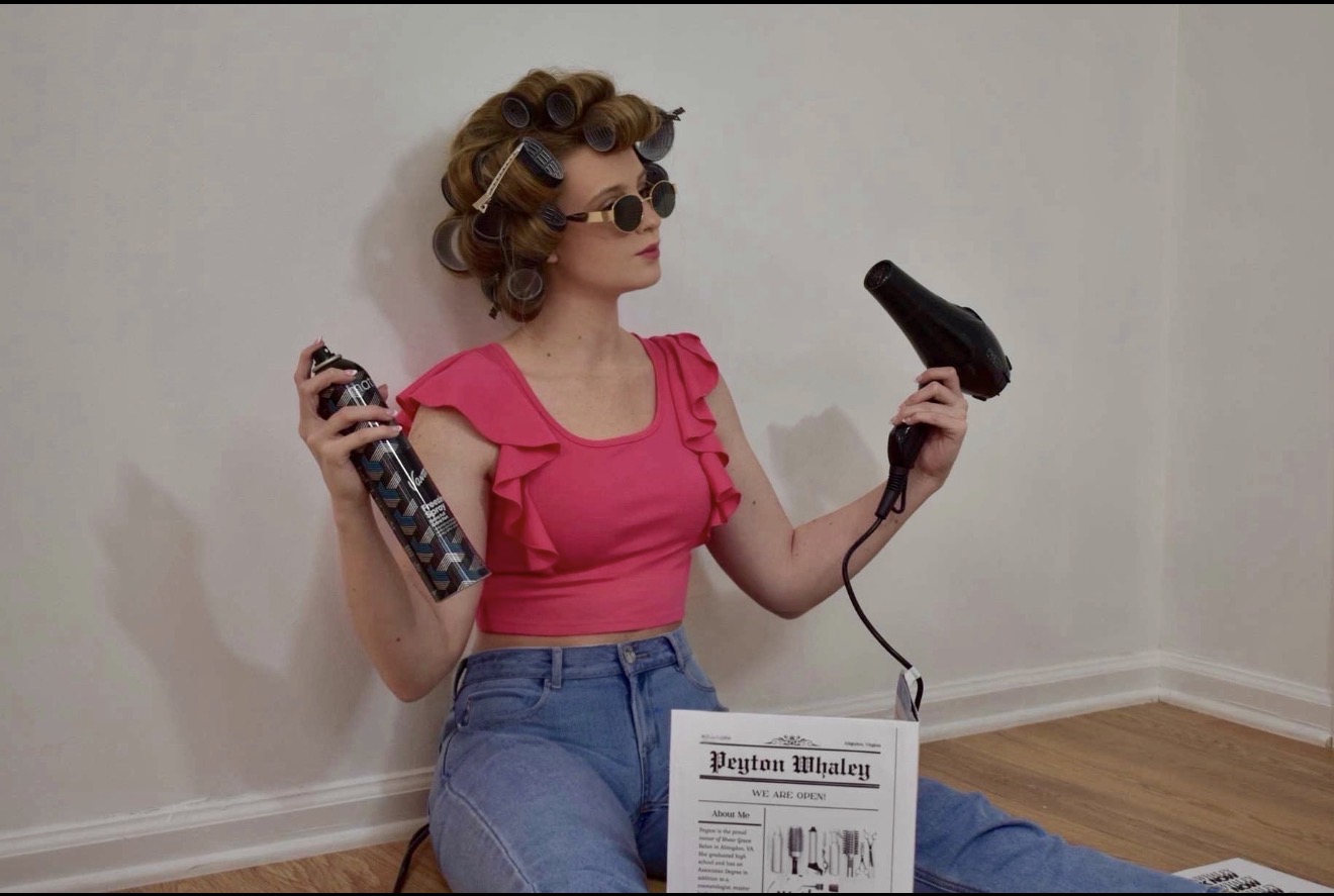 Woman with curly hair, sunglasses, pink top, and jeans sitting on the floor holding a hairdryer and a spray can, with a newspaper on her lap.