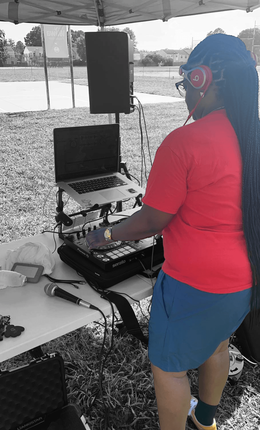 Person wearing red shirt and blue skirt with headphones operating DJ equipment outdoors under a tent.