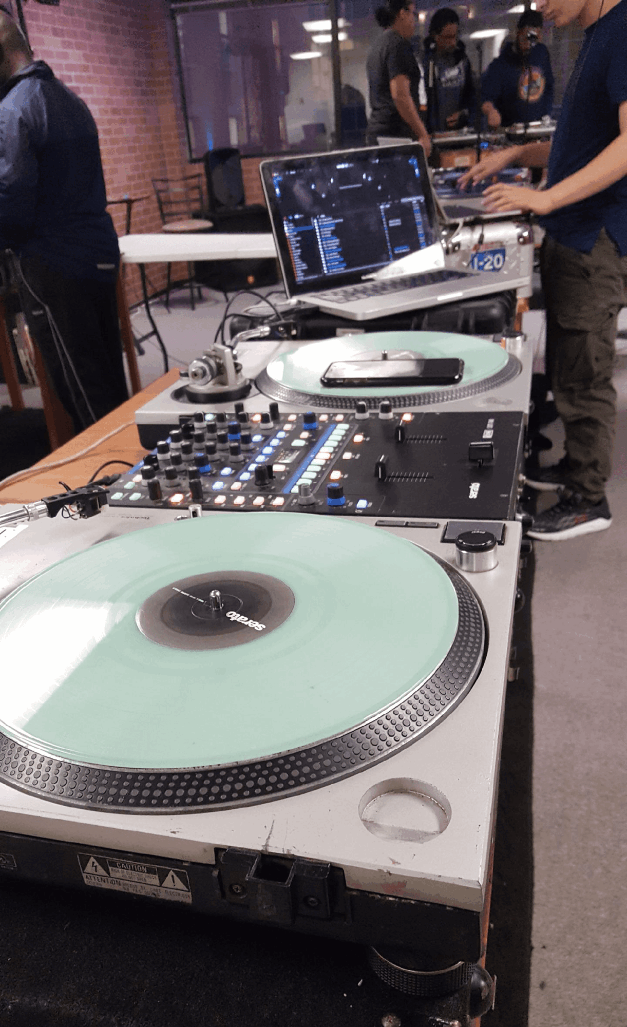 DJ equipment with turntables, mixer, and laptop on a table, people in background at an indoor event.