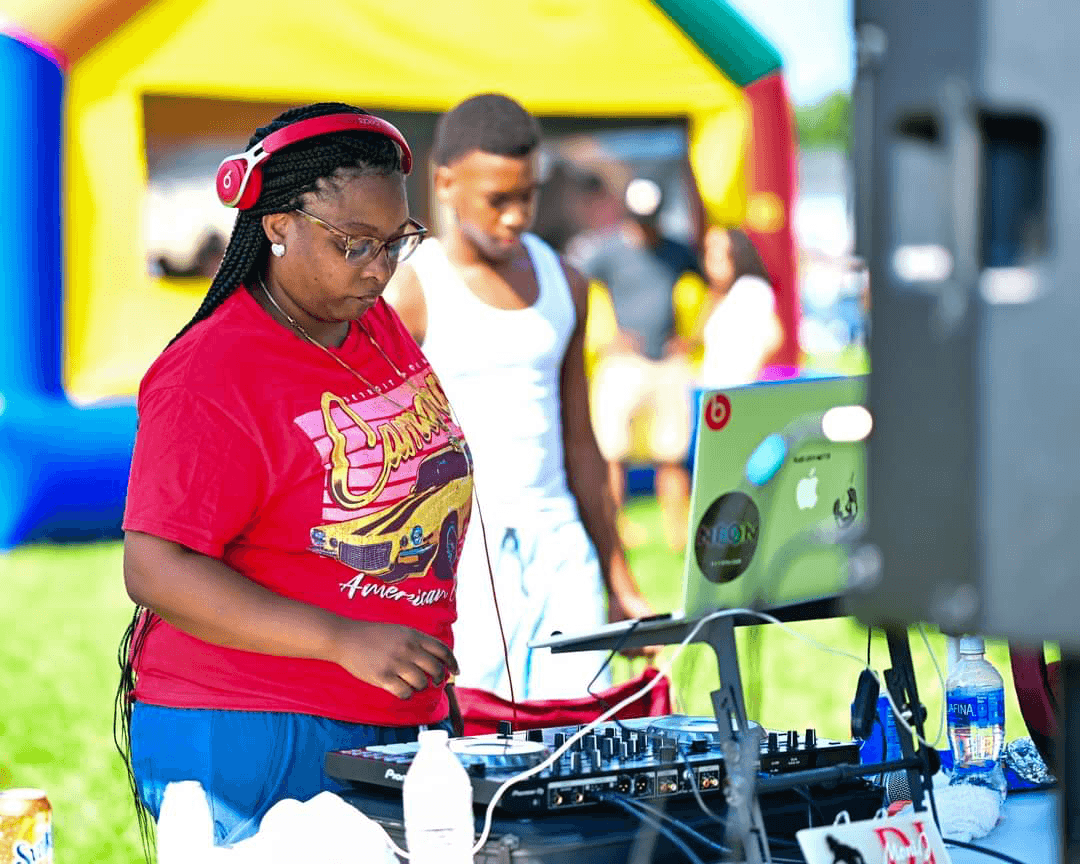 Woman wearing red shirt and headphones operating DJ equipment outdoors, with a young girl in white shirt in background.