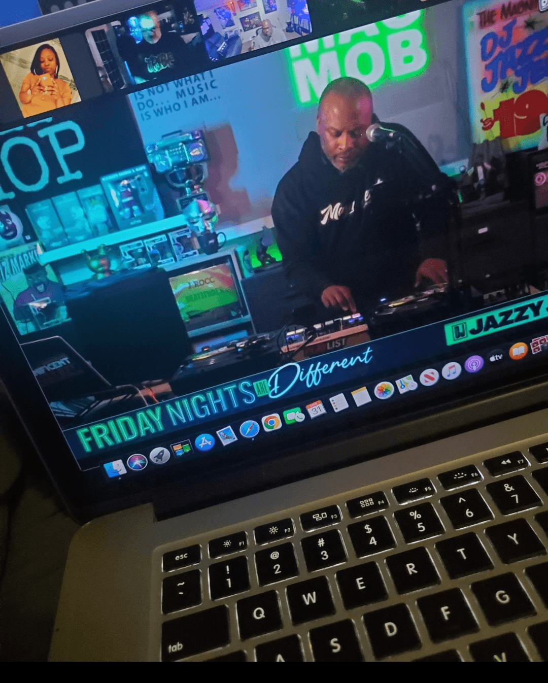 Person DJing at a colorful music venue with screens and signs, wearing a black hoodie, with a laptop in front.