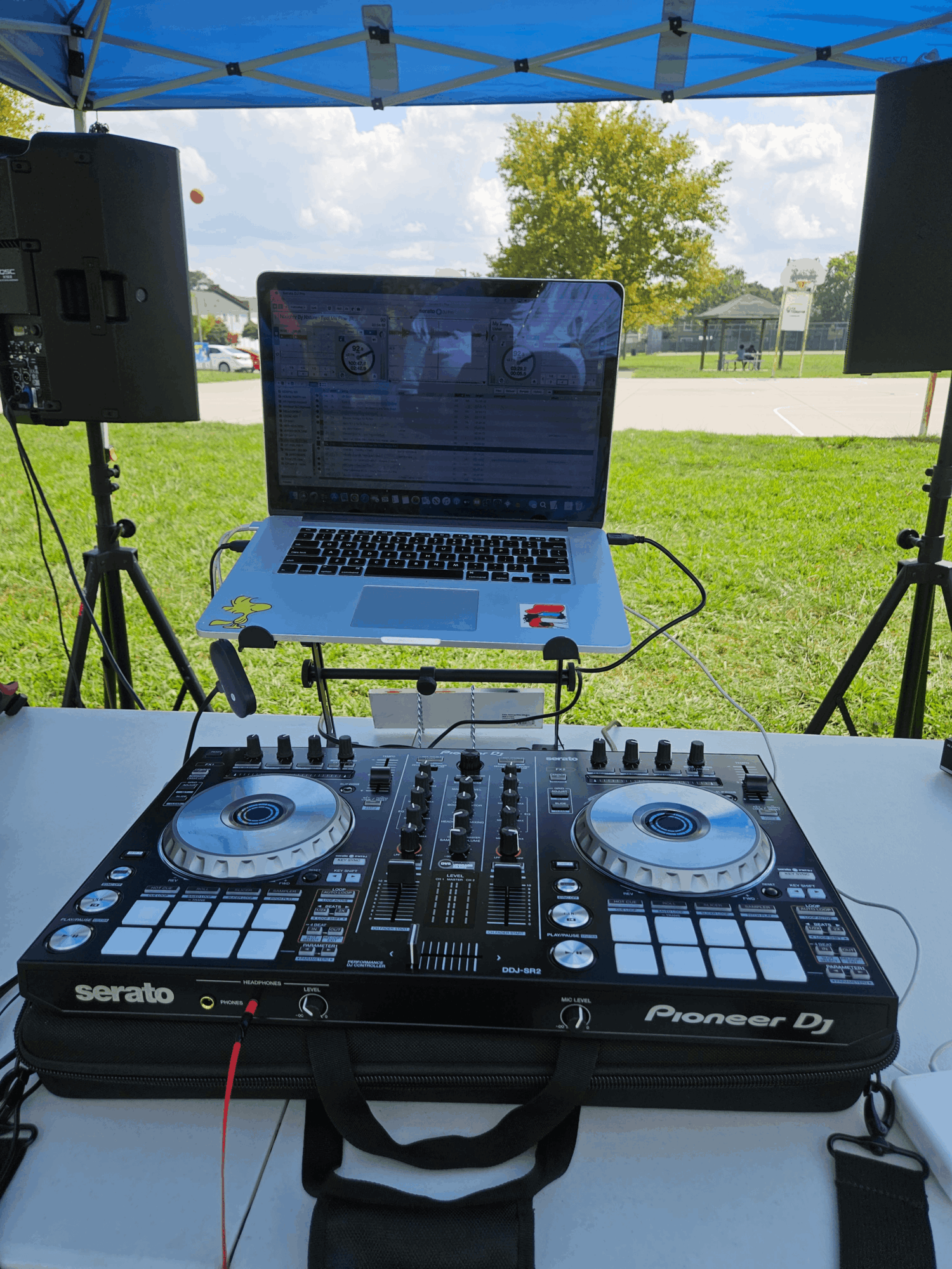 DJ equipment setup with a laptop, mixer, and speakers outdoors under a blue canopy, with grass and trees in background.
